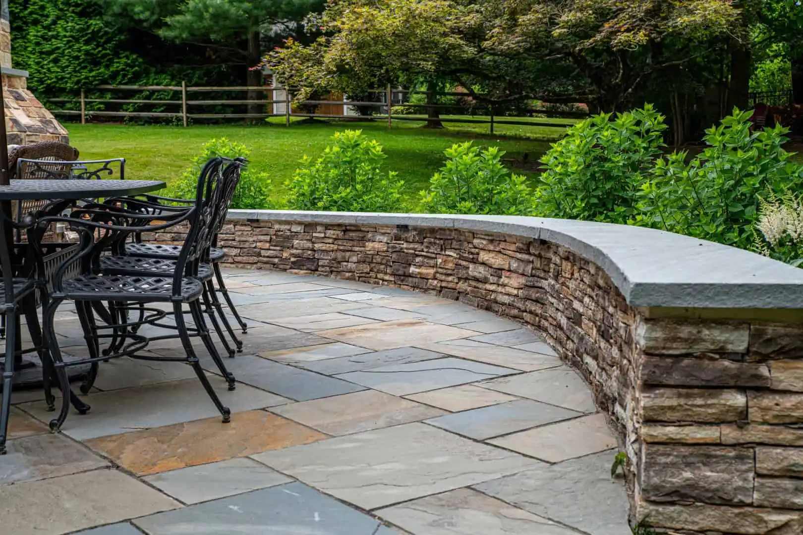 A stone patio, perfect for outdoor living in Delaware County PA, features a black metal table and chairs on the left. Bordered by a curved stone wall with lush greenery and a wooden fence behind, it creates a serene setting for relaxation.
