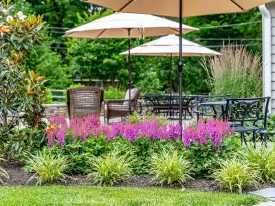 A tranquil garden with pink flowers, ornamental grasses, and a magnolia tree. Wicker and metal chairs sit alongside tables with beige umbrellas on a stone patio. Lush greenery embraces this serene outdoor living space in Delaware County, PA.