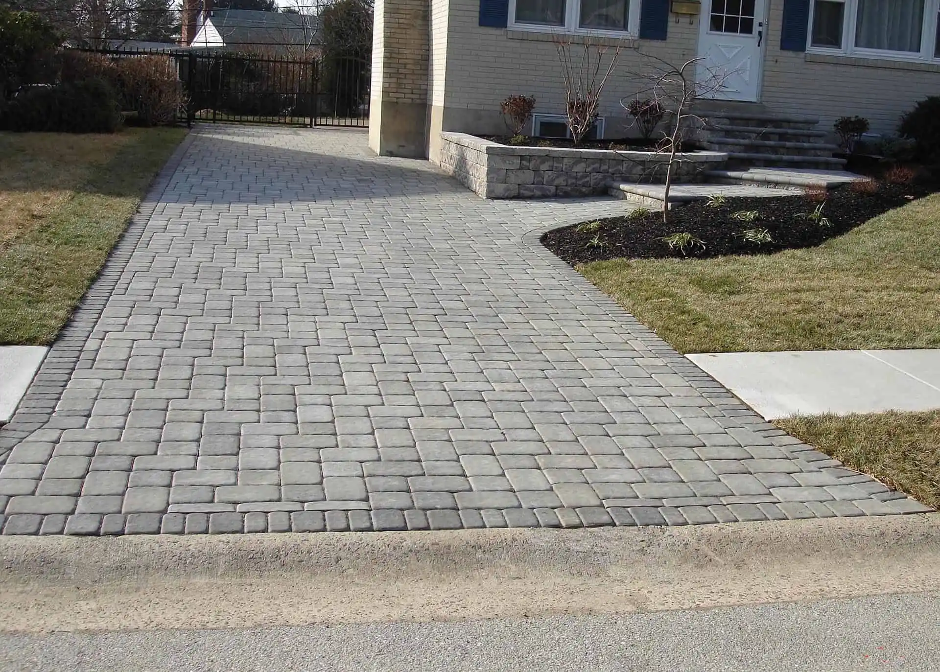 A house with a wide driveway made of rectangular gray concrete pavers, bordered by grass lawns and a small landscaped area with a young tree near the entrance.