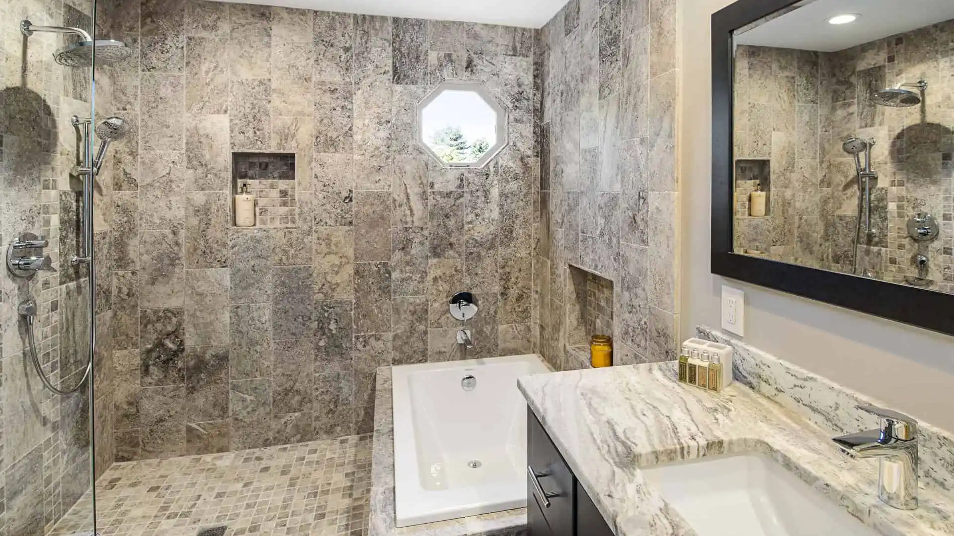 Modern bathroom with a marble countertop, rectangular sink, and large mirror. Features a step-in bathtub and a stone-tiled walk-in shower with a rainfall showerhead. A small octagonal window above the tub provides natural light echoing the beauty of outdoor living in Delaware County PA.