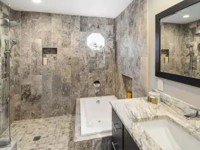 Modern bathroom with a marble countertop, rectangular sink, and large mirror. Features a step-in bathtub and a stone-tiled walk-in shower with a rainfall showerhead. A small octagonal window above the tub provides natural light echoing the beauty of outdoor living in Delaware County PA.