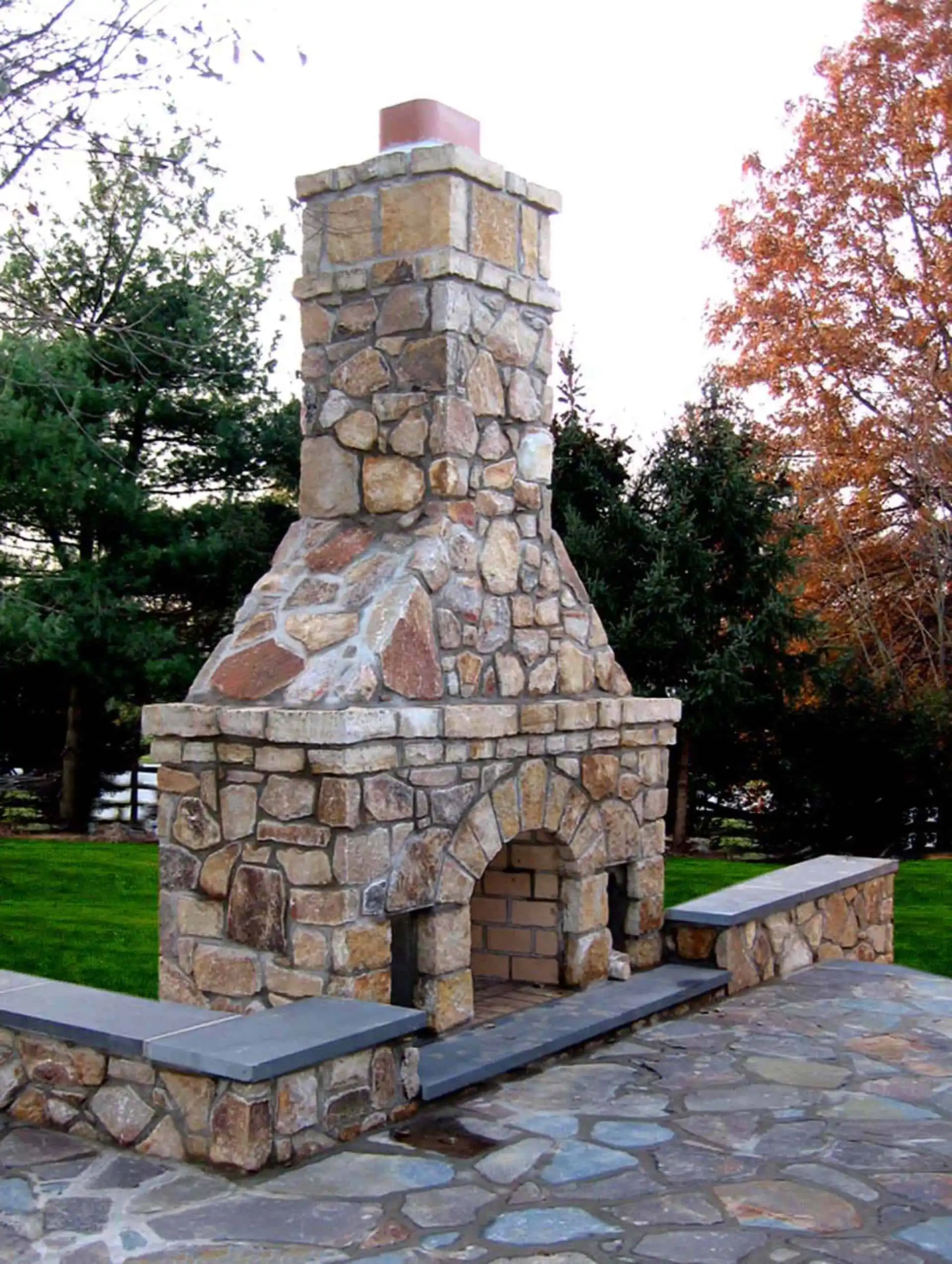 A large outdoor stone fireplace with a tall chimney, built-in seating on both sides, and surrounded by a stone patio. Green trees and autumn foliage are visible in the background.
