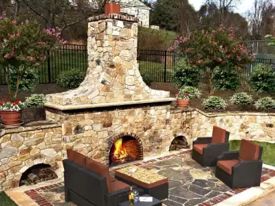 Outdoor patio featuring a stone fireplace and lit fire, perfect for outdoor living in Delaware County, PA. Surrounded by lush trees and plants, four cushioned wicker chairs encircle a rectangular stone table—expertly crafted with our hardscaping services in Delaware County, PA.