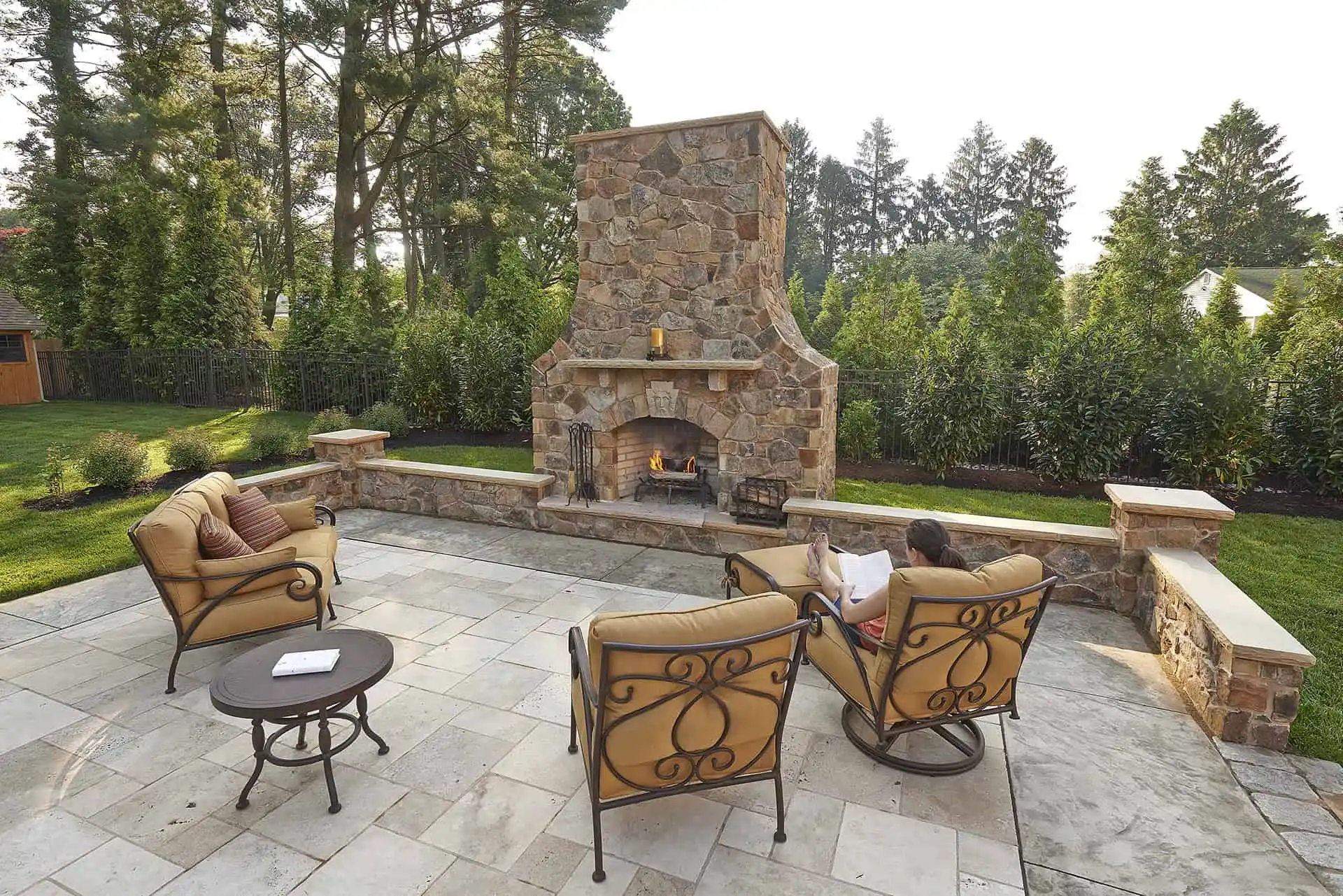 A stone outdoor fireplace on a spacious patio, surrounded by cushioned chairs and small tables. A person sits in one chair reading, with trees and greenery providing a scenic backdrop.