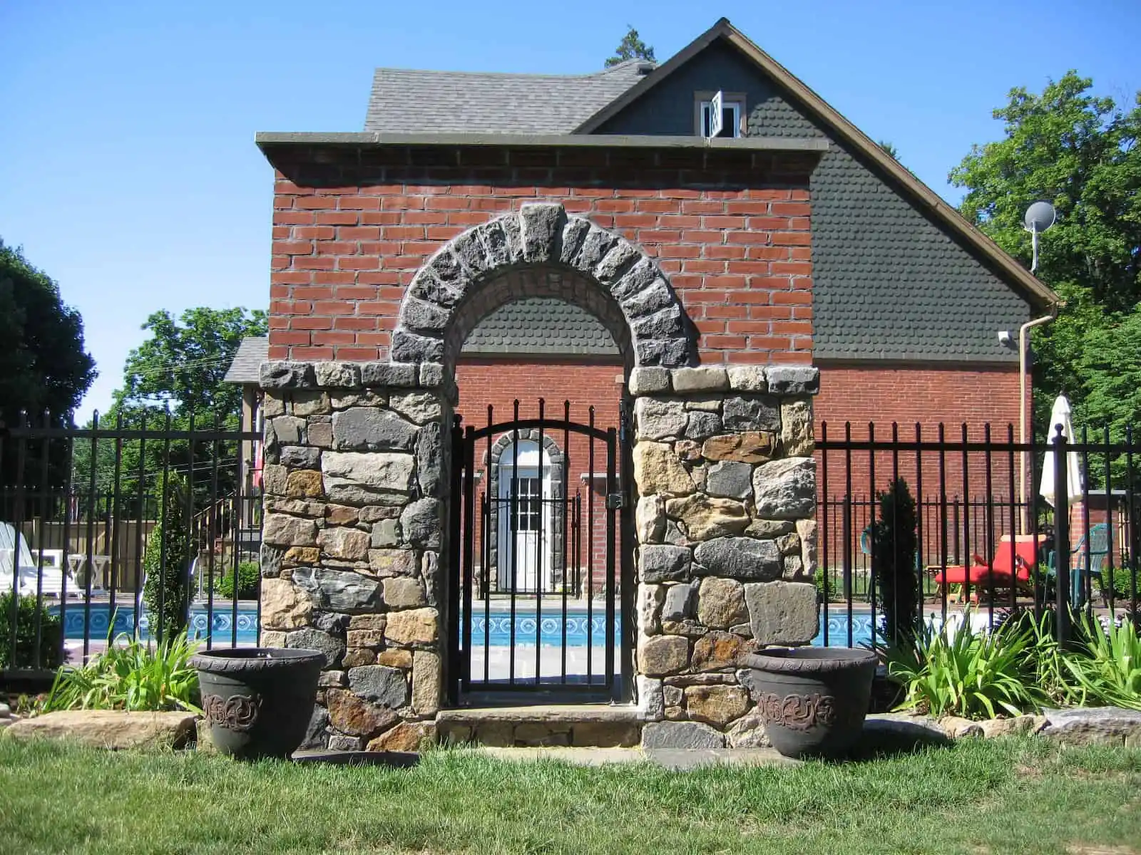 A stone and brick archway with a black metal gate stands in front of a swimming pool, with a brick house and green lawn in the background. Two large planters are placed on either side of the archway.