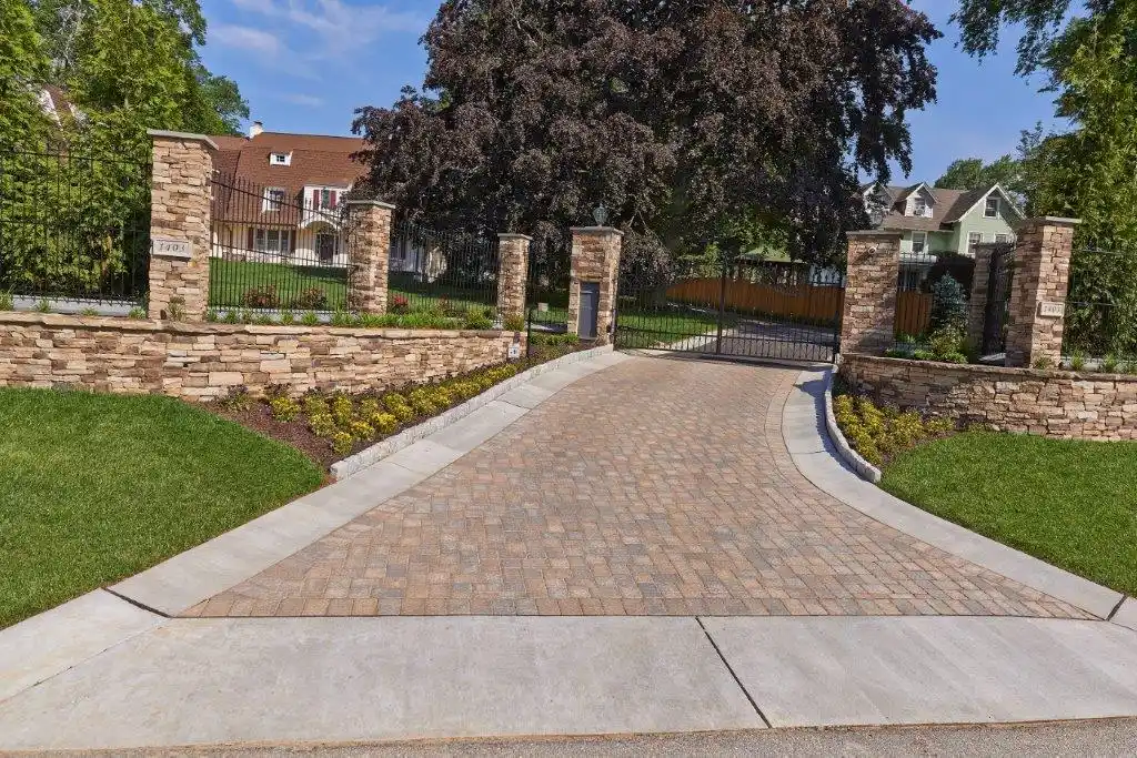 A paved driveway leads to a large gate surrounded by stone pillars, showcasing expertly done hardscaping services in Delaware County, PA. The entrance, flanked by manicured lawns and shrubs, hints at the outdoor living elegance with a house partially visible behind the lush trees.