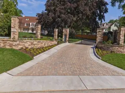 A paved driveway leads to a large gate surrounded by stone pillars, showcasing expertly done hardscaping services in Delaware County, PA. The entrance, flanked by manicured lawns and shrubs, hints at the outdoor living elegance with a house partially visible behind the lush trees.