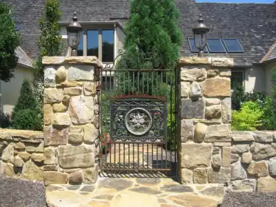 A wrought iron gate with an intricate design, flanked by two stone pillars topped with lanterns, offers a stunning entrance crafted through expert hardscaping services in Delaware County PA. As you pass through, a path winds towards a house with gray tiles and skylights, nestled among lush greenery and tall trees.