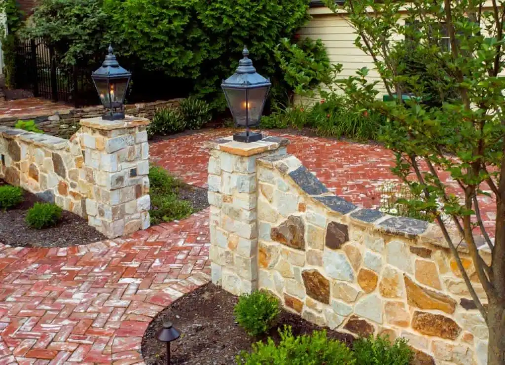 A stone and brick pathway with lamp posts leads to a curved wall lined with greenery, showcasing expert landscaping services in Delaware County, PA. A small tree and decorative shrubs border the walkway, enhanced by soft outdoor lighting for a tranquil outdoor living experience.