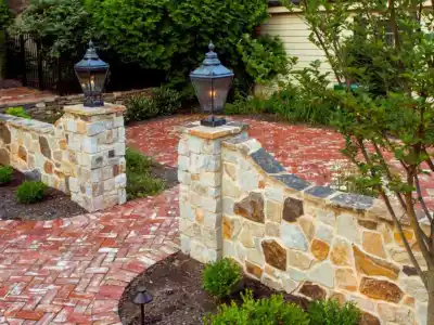 A stone and brick pathway with lamp posts leads to a curved wall lined with greenery, showcasing expert landscaping services in Delaware County, PA. A small tree and decorative shrubs border the walkway, enhanced by soft outdoor lighting for a tranquil outdoor living experience.