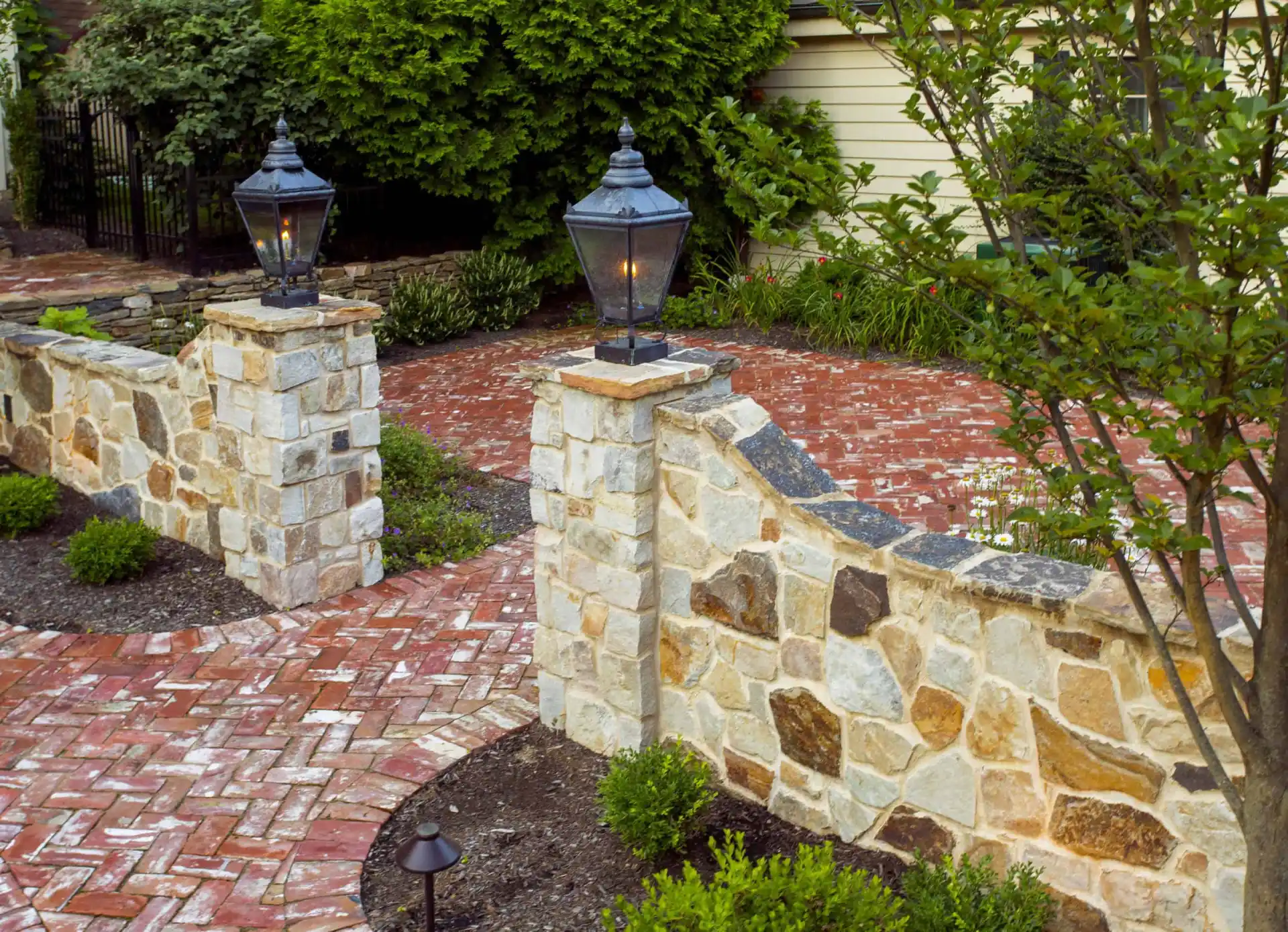 A stone and brick garden wall with two lantern-topped pillars borders a brick walkway. Small bushes and plants line the path, with trees and a house visible in the background.