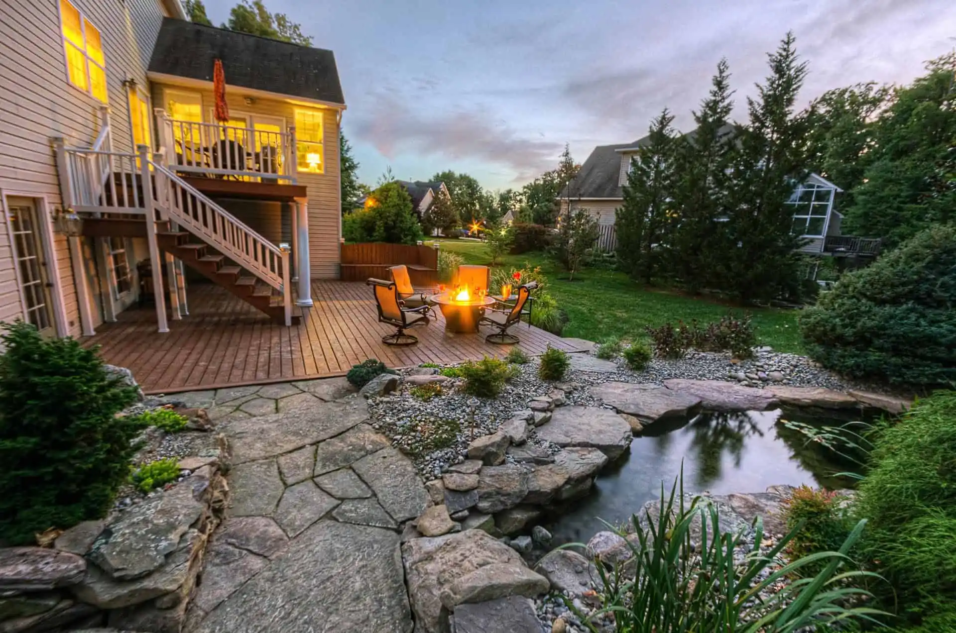 Backyard patio with a fire pit surrounded by chairs, stone pathway, small pond with rocks, plants, and a wooden deck attached to a house at dusk. Lit windows and green lawn create a cozy atmosphere.