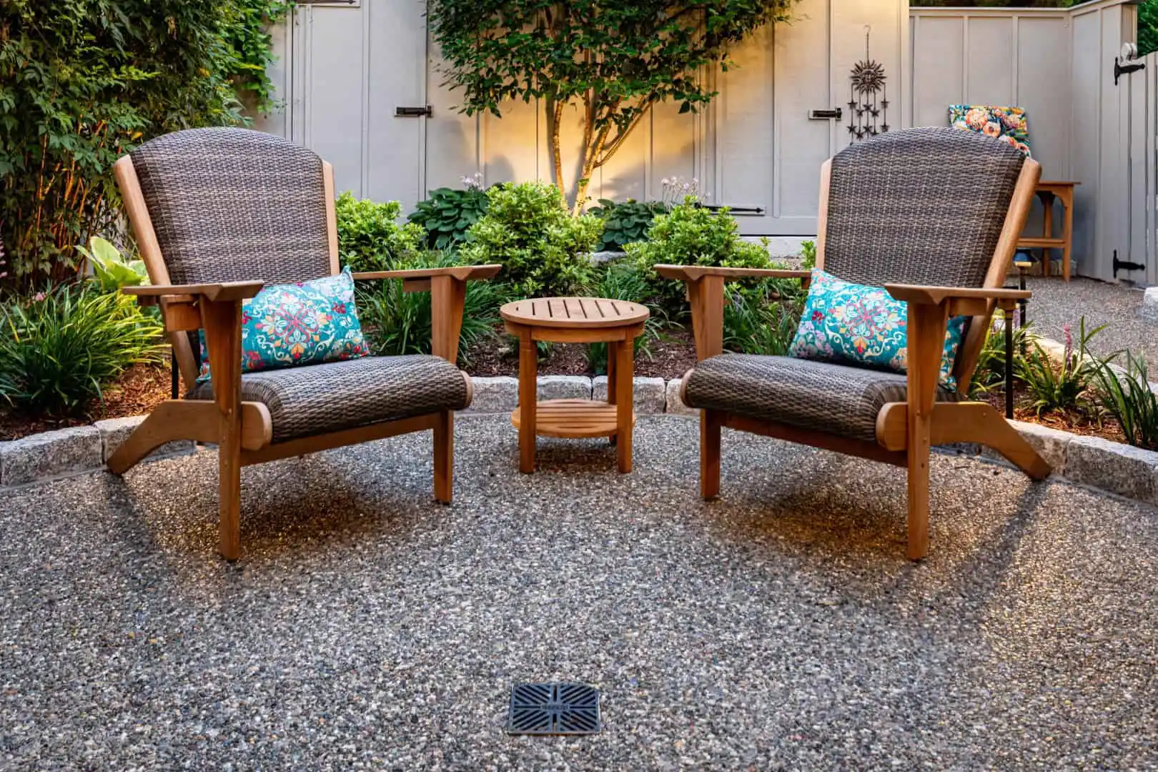 Two wooden chairs with woven backs sit on a pebble patio, each adorned with colorful cushions. A small round wooden table is between them. The serene setting showcases expert hardscaping services in Delaware County PA, framed by lush greenery and a softly lit fence.