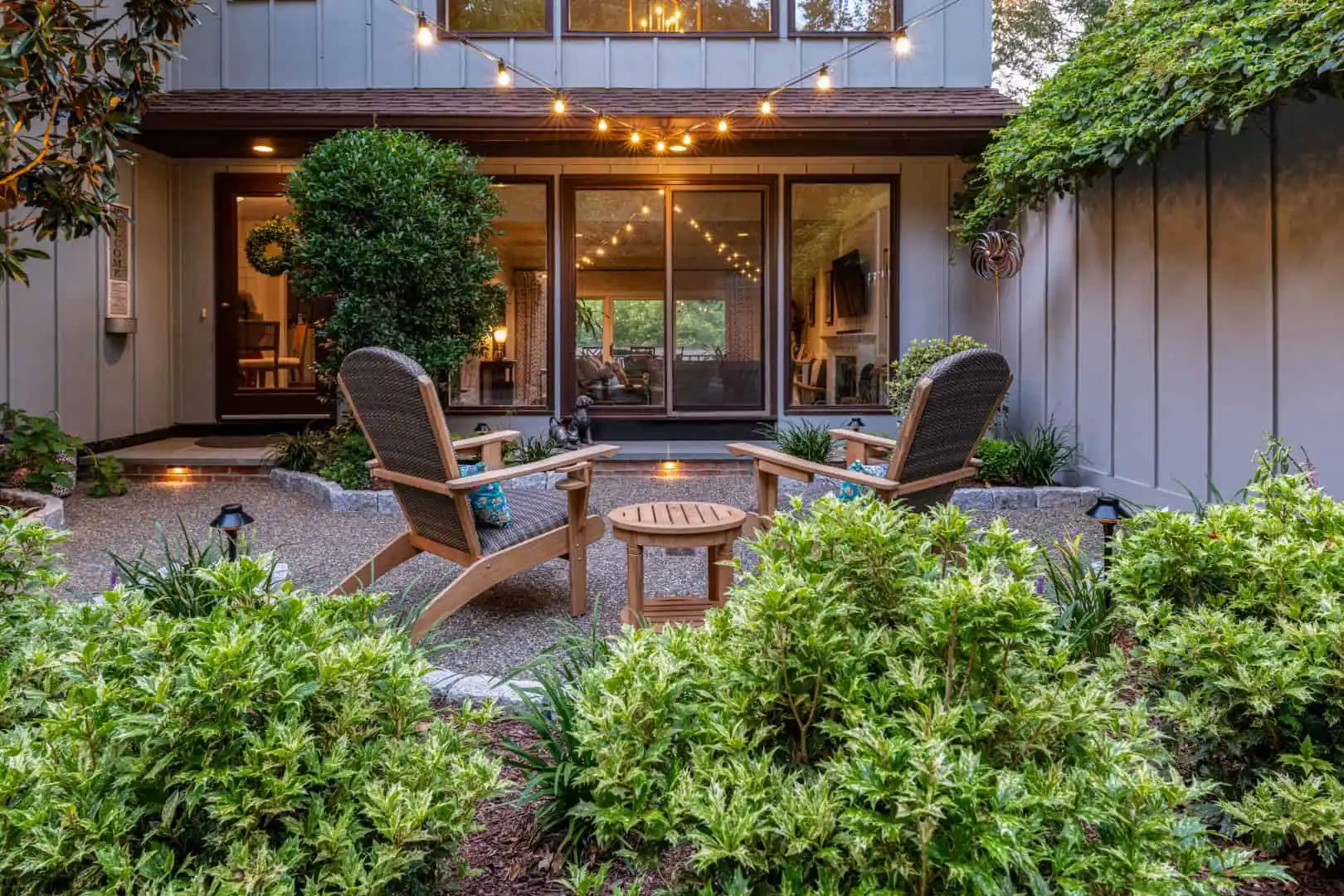 A cozy patio with four wooden chairs and a round table, lit by string lights, offers the perfect spot for outdoor living in Delaware County, PA. Surrounded by lush greenery and shrubs, it complements the house's large glass doors that reveal a warmly lit interior.