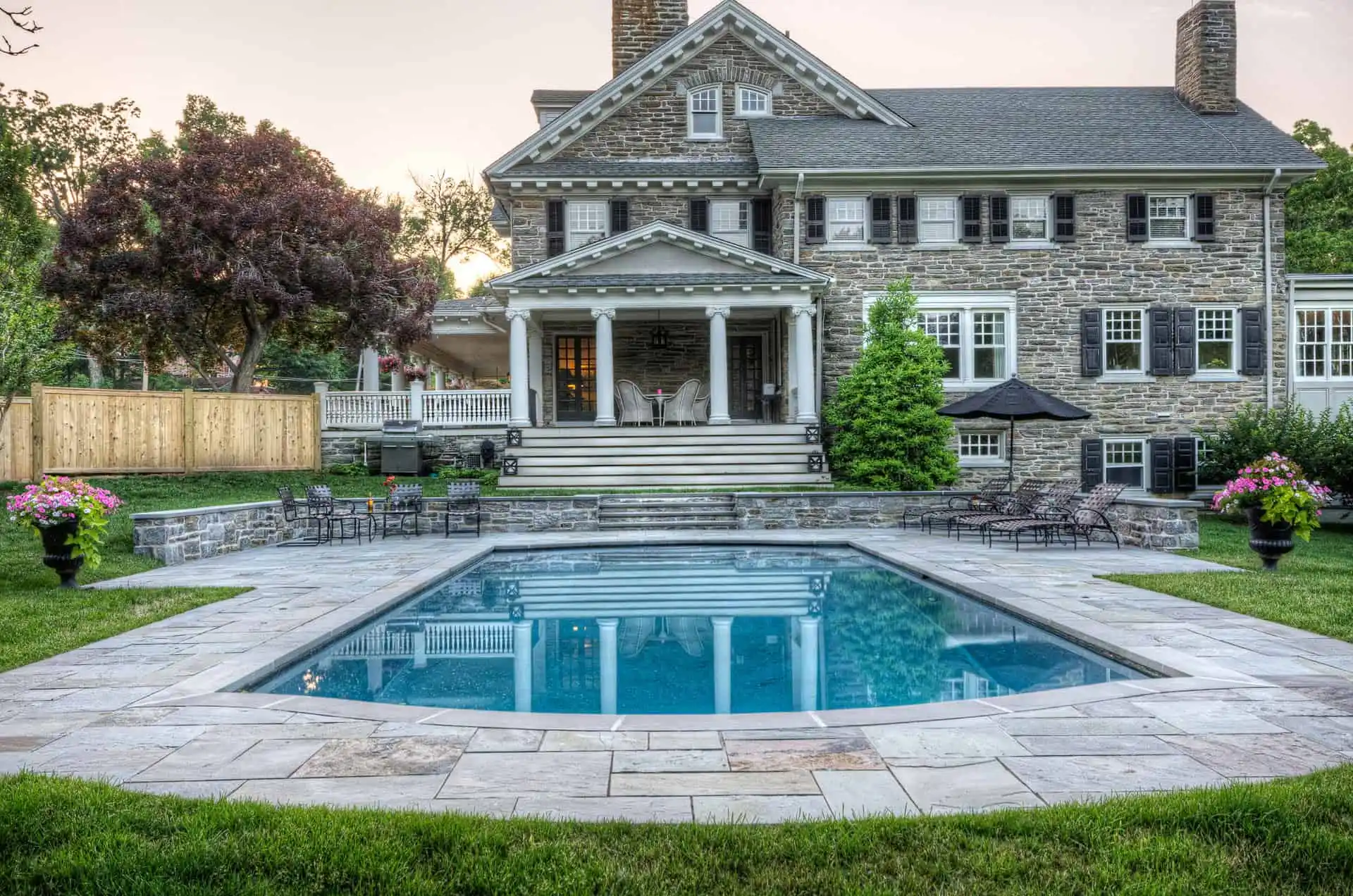 A large stone house with white trim features a backyard swimming pool, surrounded by a stone patio, green grass, potted flowers, and outdoor seating. The home's columns and landscaping add elegance.