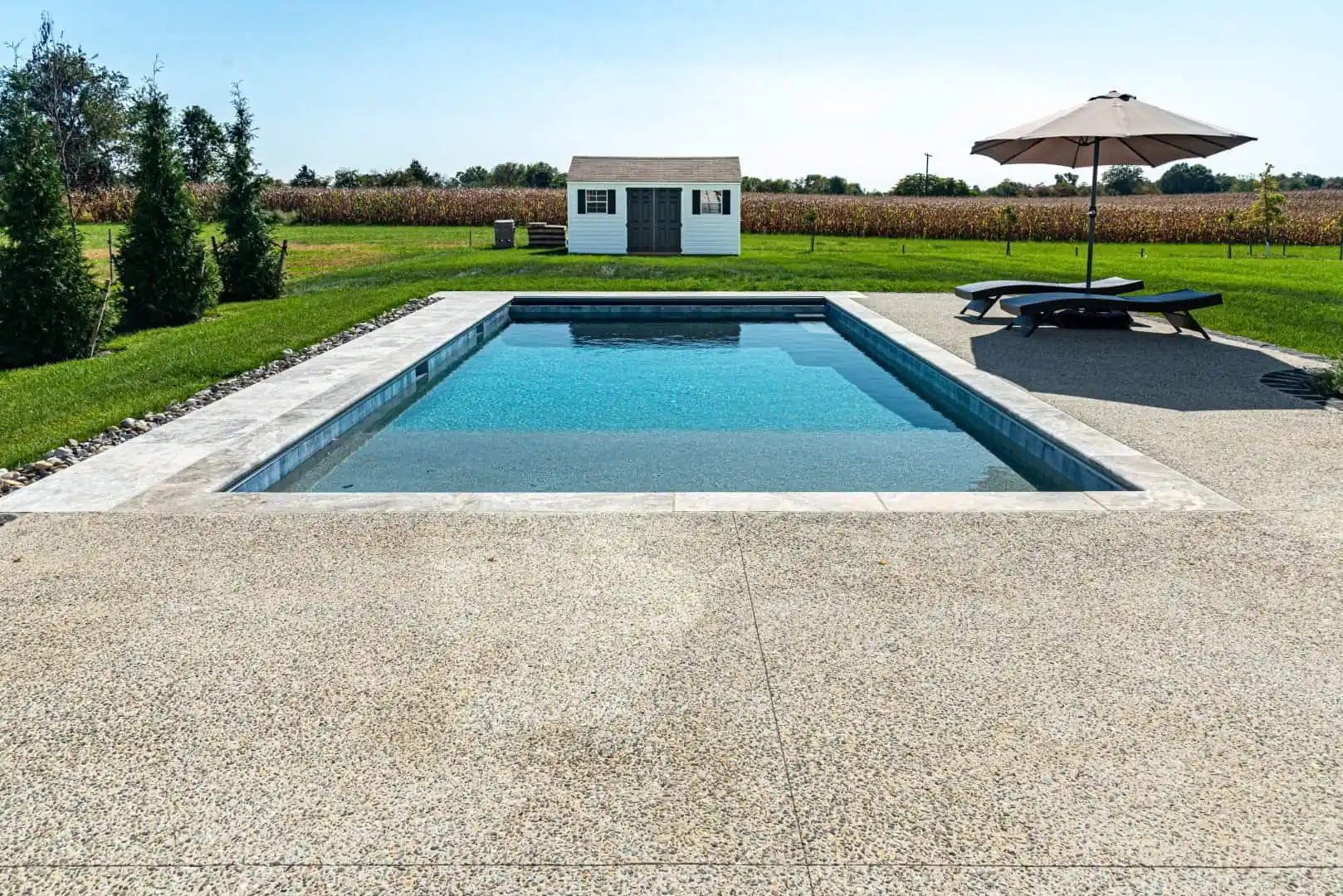 A rectangular swimming pool with clear blue water is surrounded by a concrete deck, expertly crafted through hardscaping services in Delaware County, PA. Two lounge chairs sit under a large umbrella on the right. In the background, there is a small white shed and a field with trees along the horizon.
