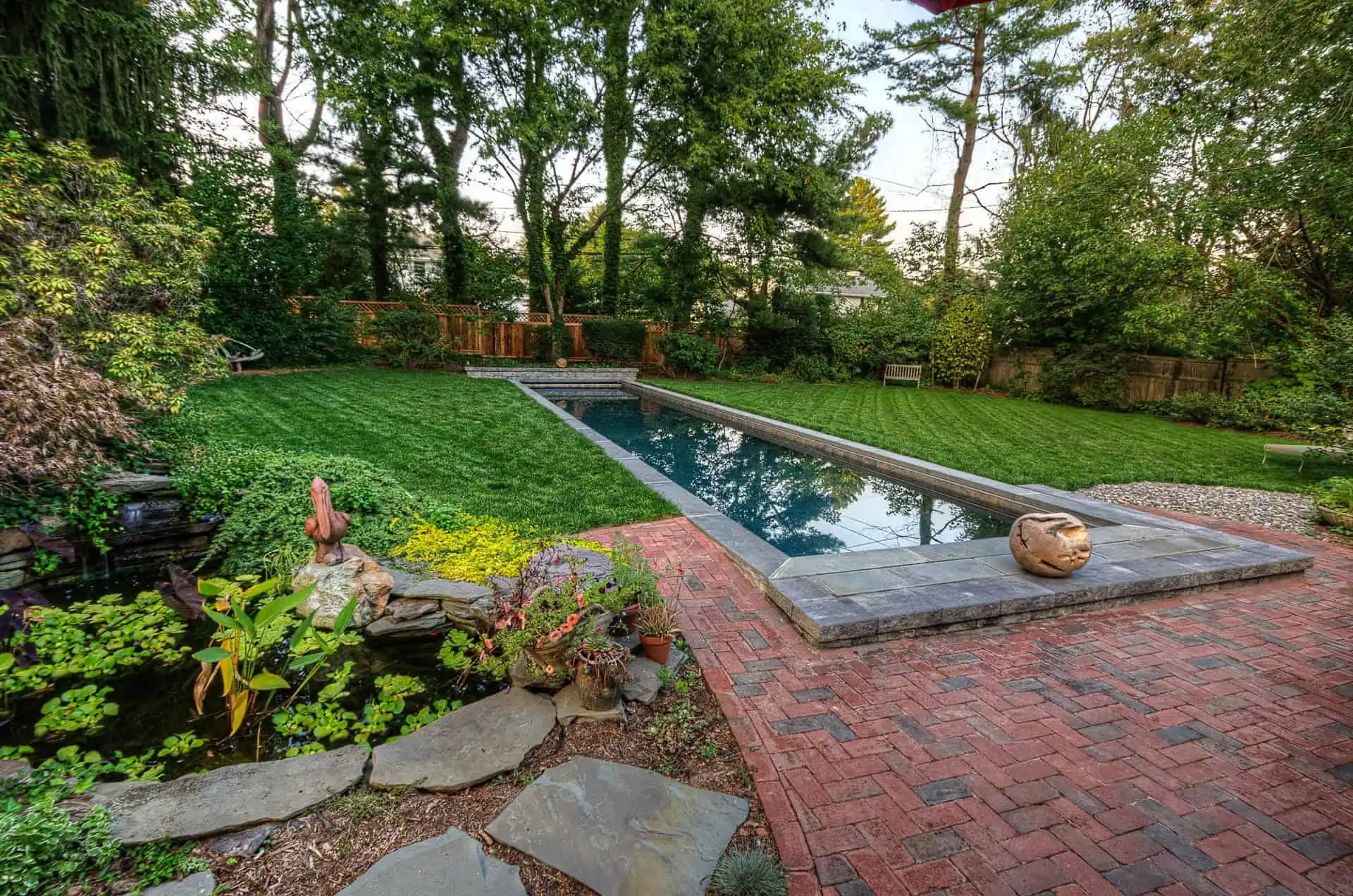 A backyard with a long rectangular pool, surrounded by green grass, trees, and shrubs. Brick paving lines the poolside, with garden statues and plants near a small pond in the foreground.