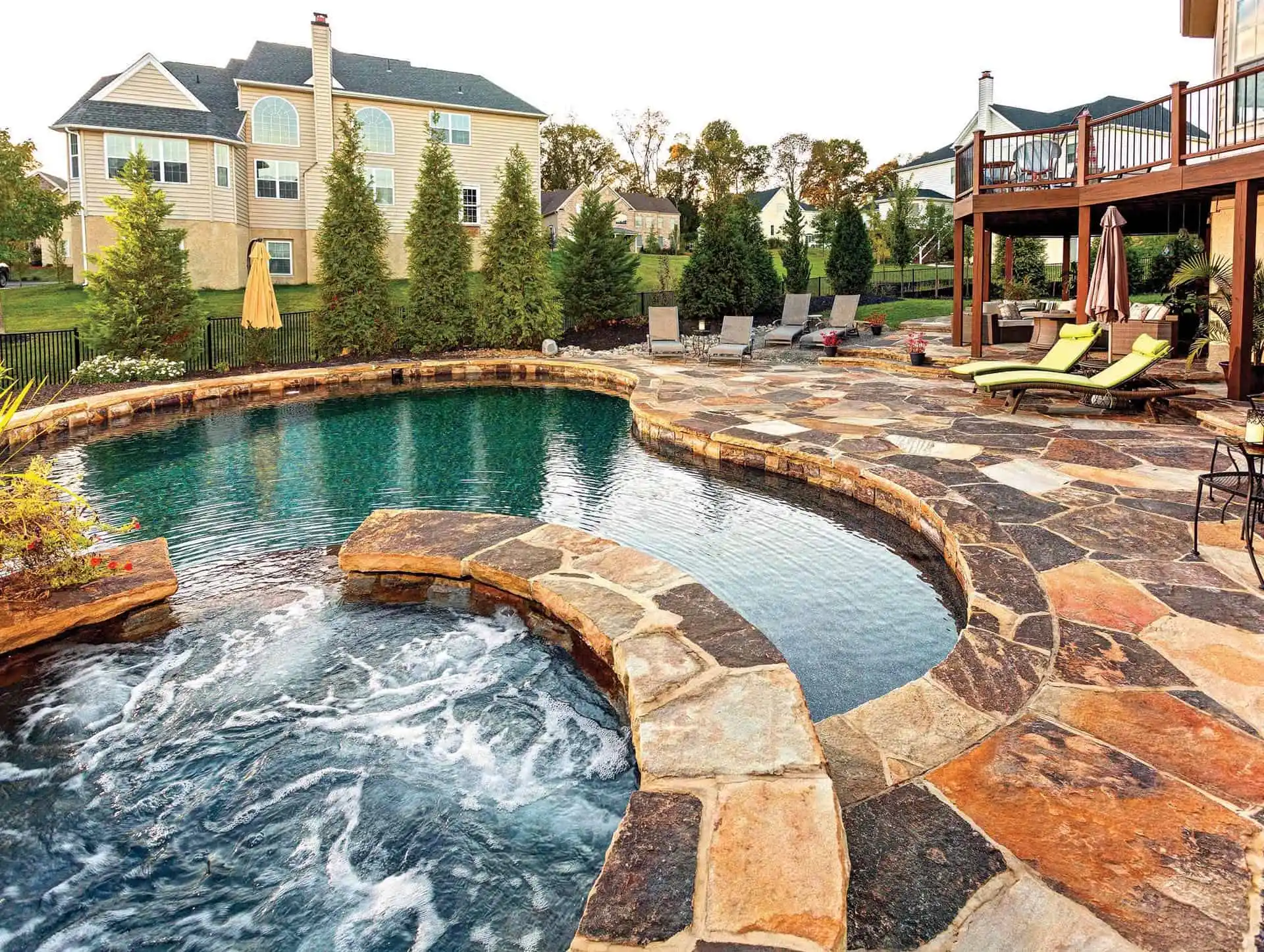 A backyard with a stone patio features a circular spa connected to a swimming pool. Several lounge chairs and umbrellas are set up, with trees and large houses visible in the background.