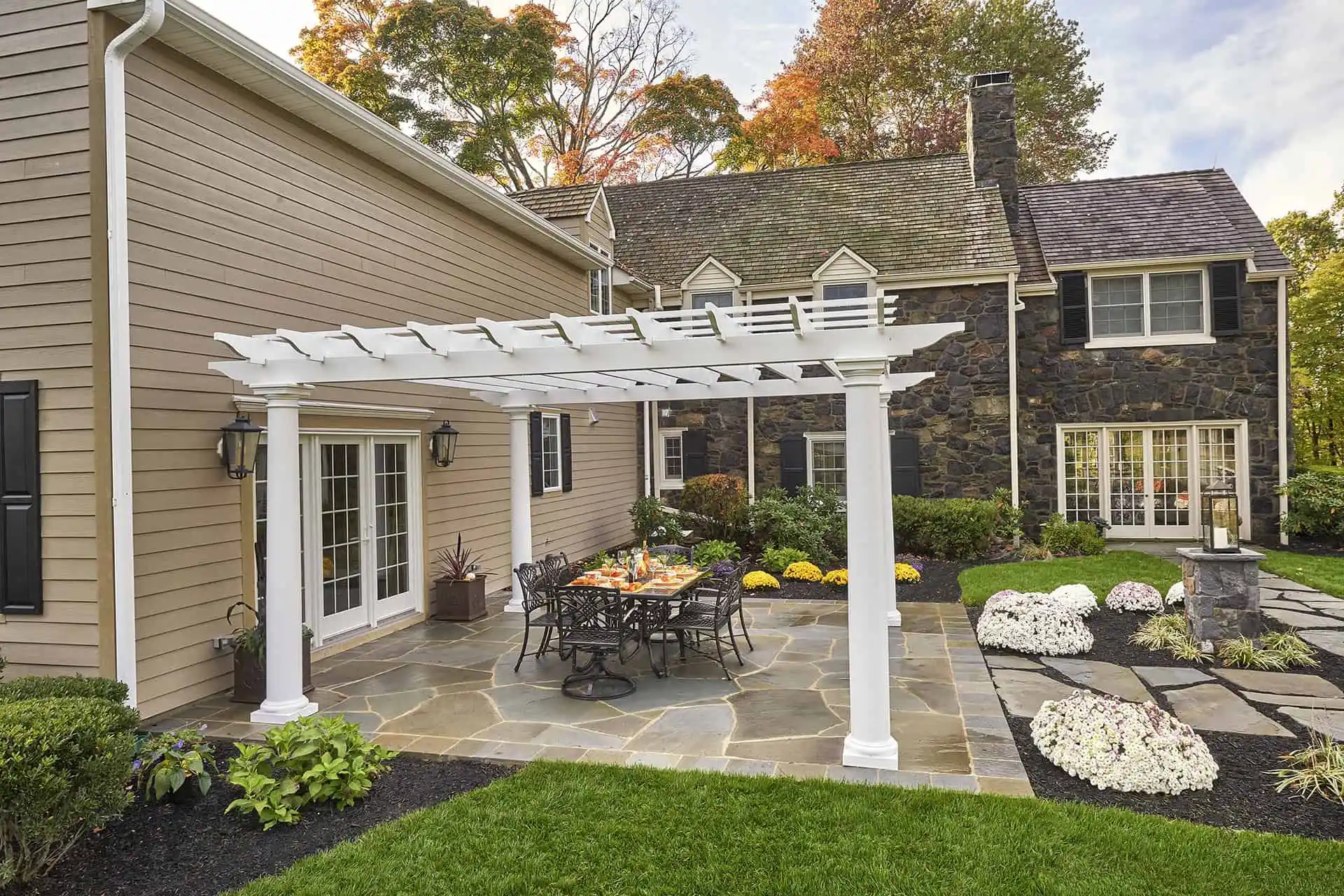 A stone patio with a white pergola, outdoor dining table, and chairs sits next to a beige house with black shutters and a stone extension, surrounded by landscaped gardens and flower beds.