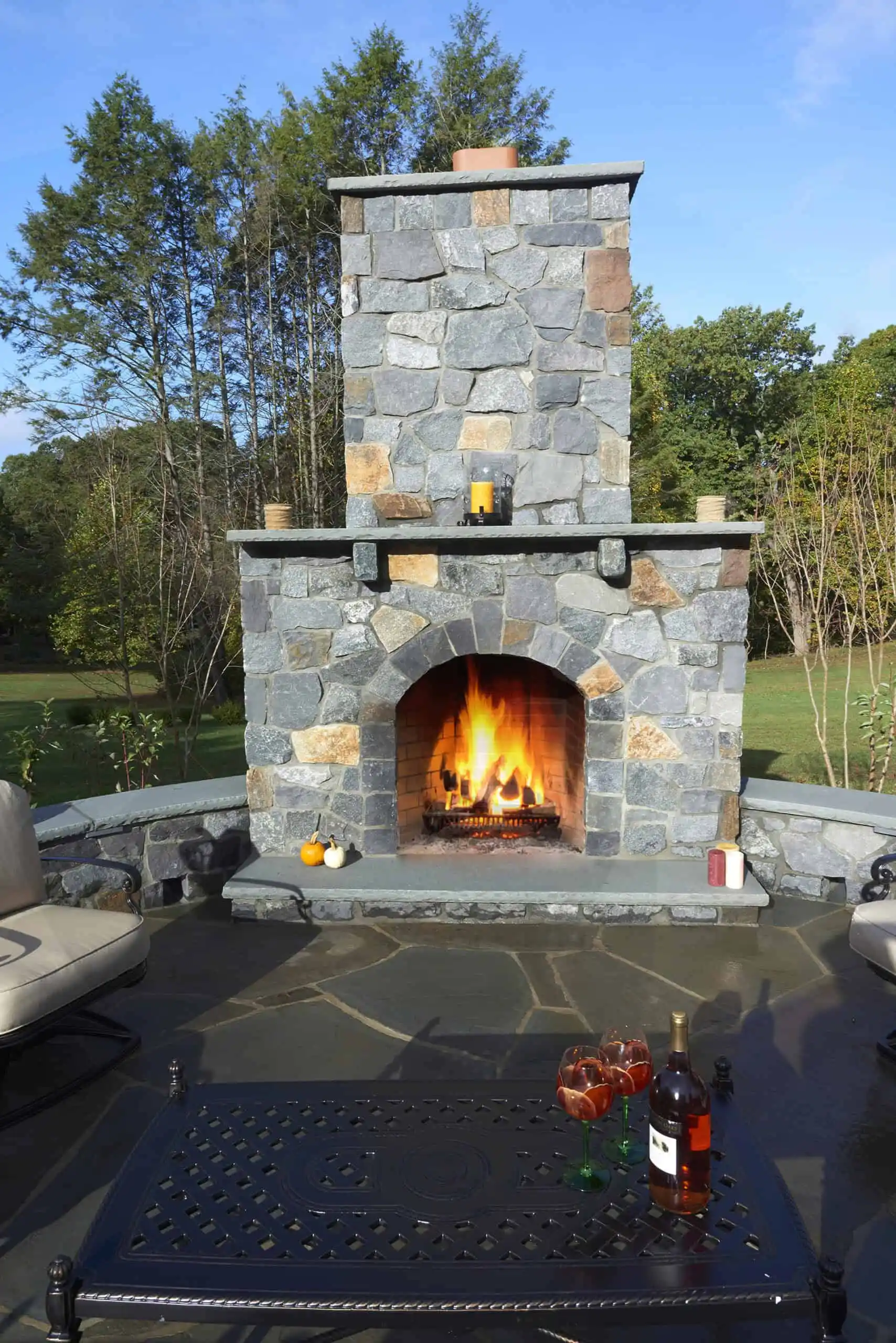 An outdoor stone fireplace with a fire burning, surrounded by patio seating. A black table in front holds glasses with red drinks and a bottle. Green trees and grass are visible in the background.