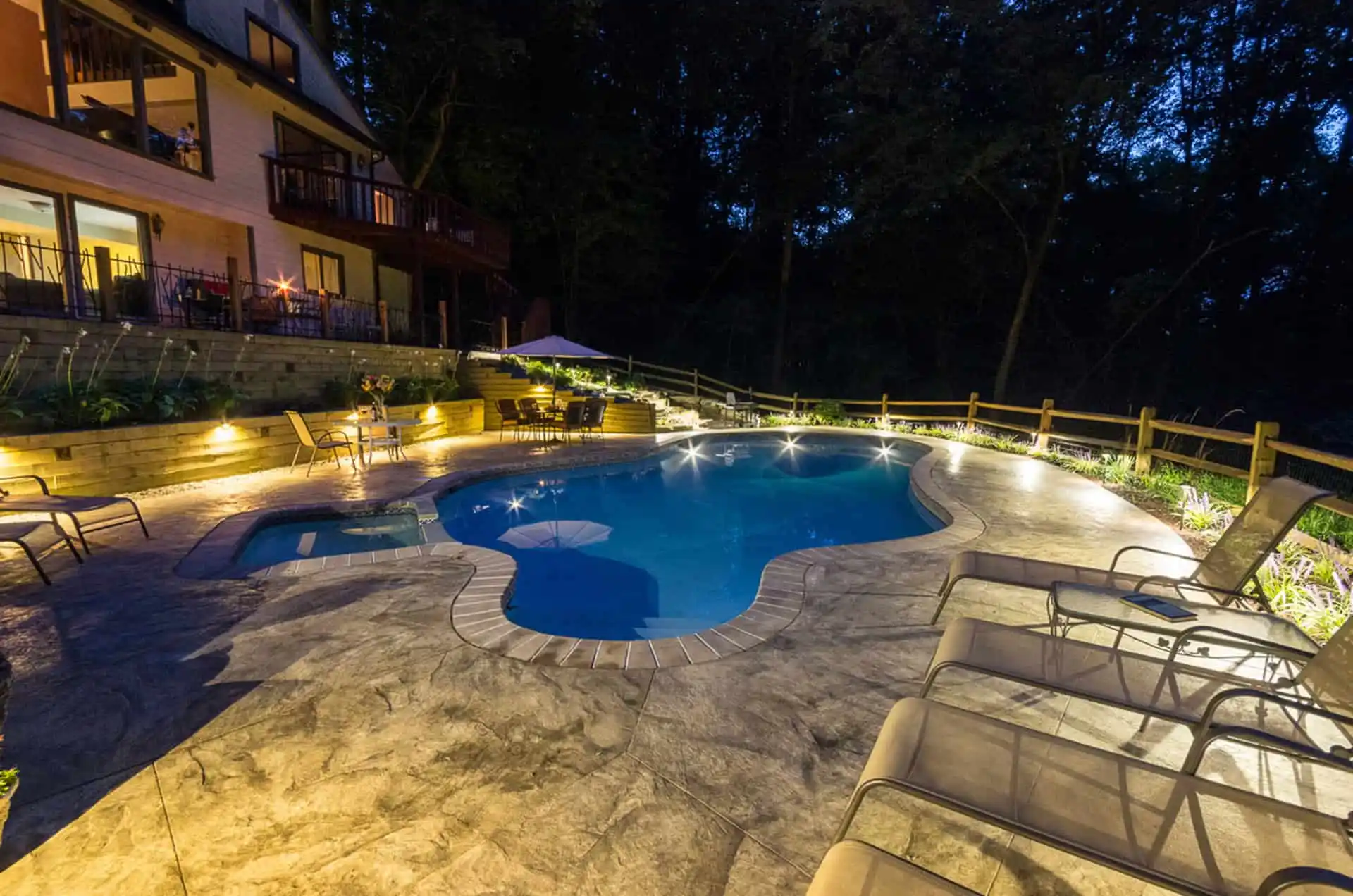 A backyard swimming pool is lit up at night, surrounded by lounge chairs, umbrellas, and landscaping, with a house and a wooded area in the background.