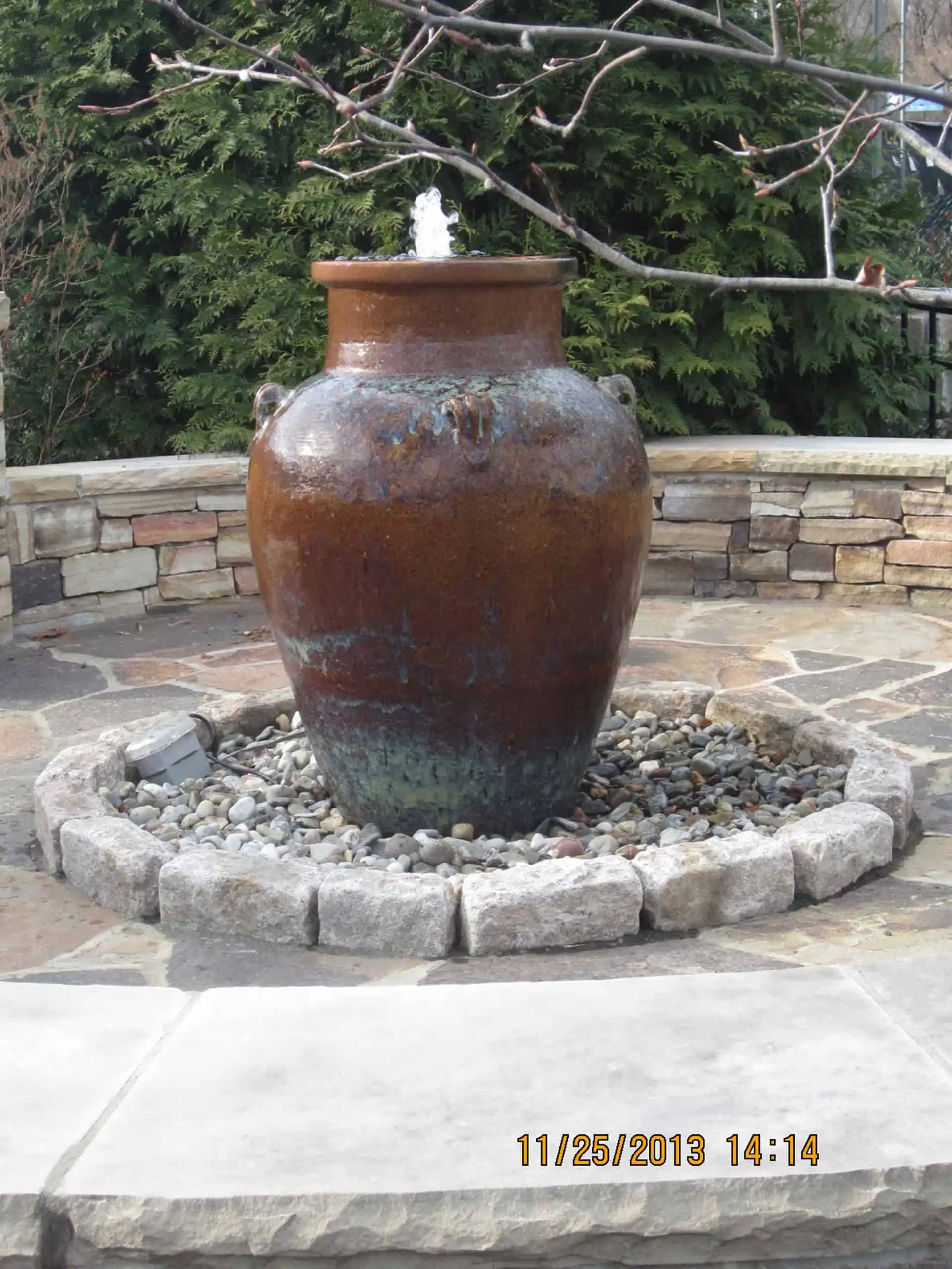 A large brown ceramic urn fountain with water bubbling from the top, surrounded by rocks and a stone border, set against a stone wall and greenery.