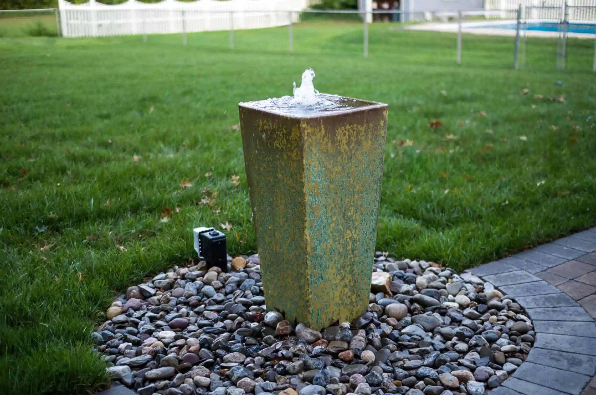 A square, greenish stone fountain stands on a bed of smooth rocks in a grassy backyard, with a small stream of water bubbling from the top. A paved walkway and white fence are visible in the background.