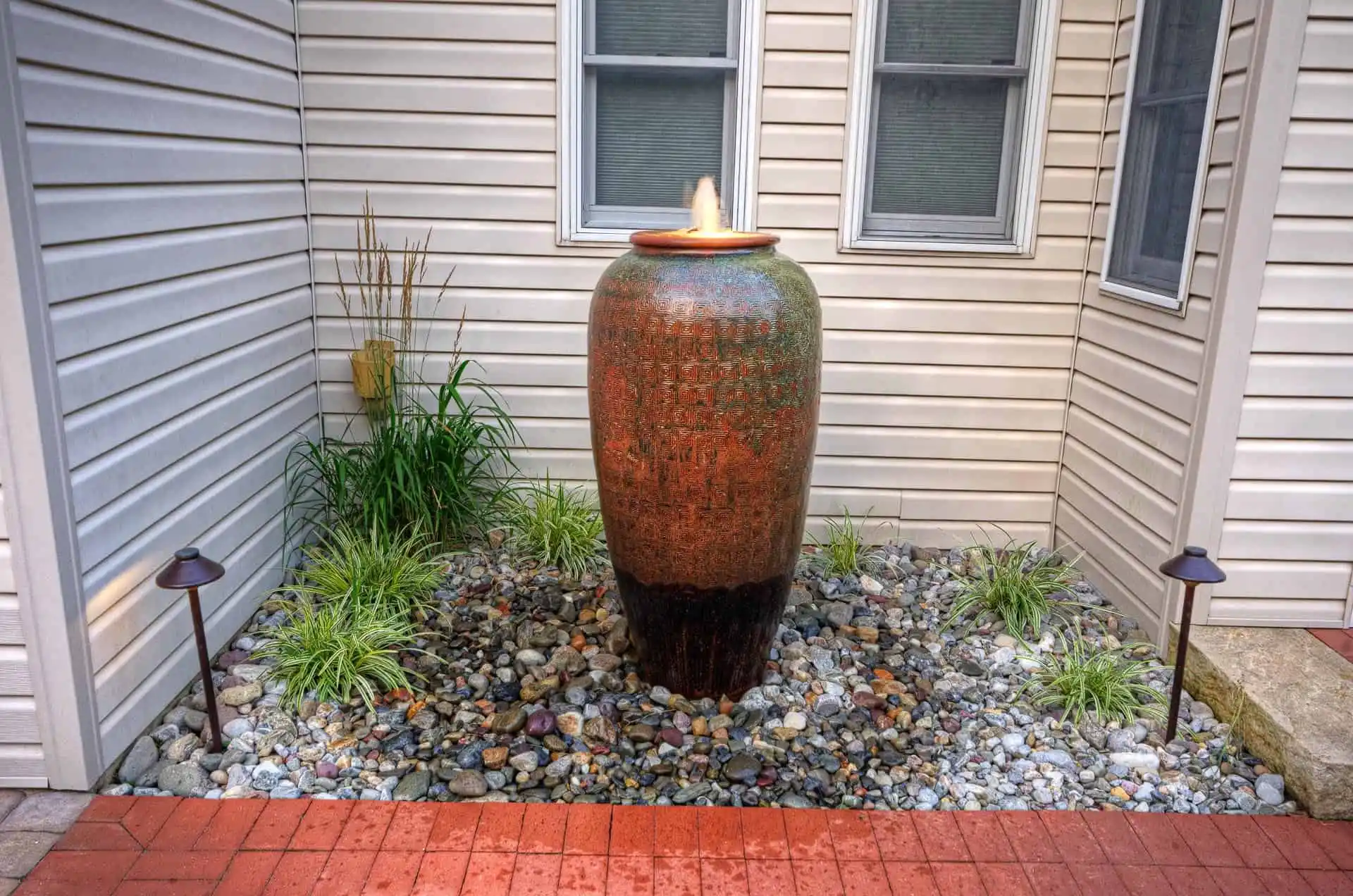 A tall ceramic urn fountain sits on a bed of rocks surrounded by green plants in a corner of a house exterior with beige siding and two windows. Two small lights stand on either side of the urn.