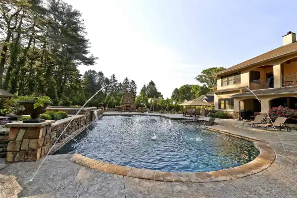 Luxurious backyard with a swimming pool featuring water jets, surrounded by stone decking, lush landscaping, lounge chairs, and a large two-story house with balconies in the background.