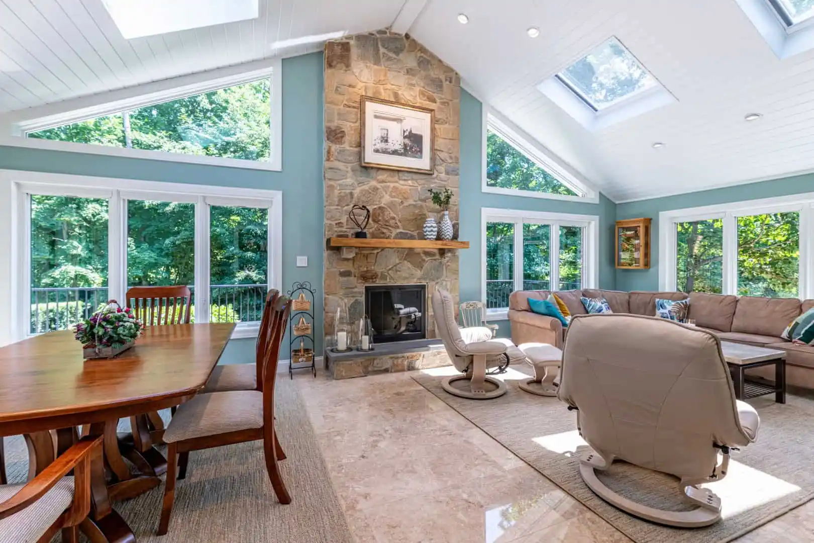 Bright sunroom with large windows and skylights, perfect for enjoying nature views. Features a stone fireplace, beige recliners, a sectional sofa, and a wooden dining table. Embrace outdoor living Delaware County PA style with potted plants that bring the beauty of your garden inside.