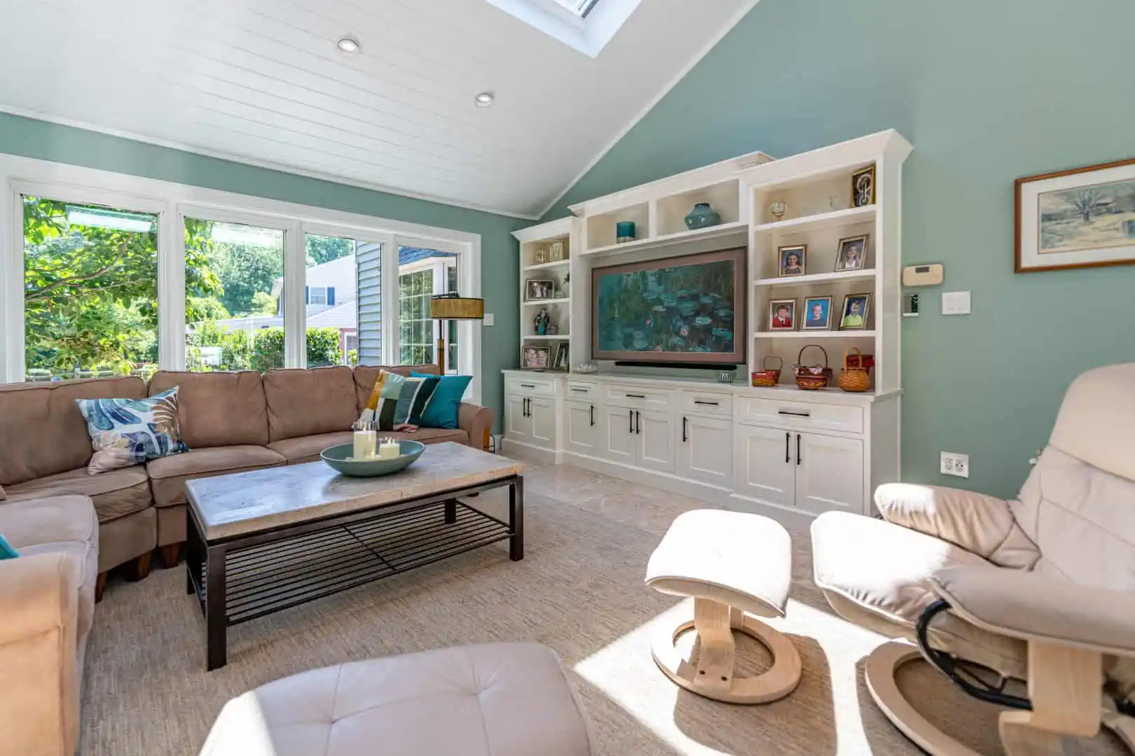 Bright living room with a large beige sectional sofa and a white entertainment center featuring TV and decor accents under a skylight. Windows and a wooden coffee table add charm, while light blue walls and a tan recliner enhance the cozy ambiance, echoing outdoor living vibes of Delaware County PA.