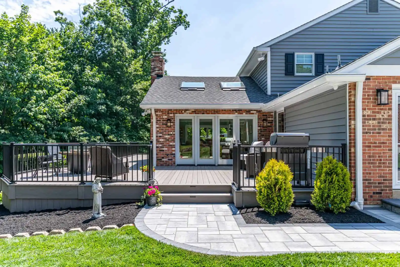 The image showcases a house with a spacious backyard patio, ideal for outdoor living in Delaware County PA. Featuring a gray deck with railings, potted plants, and a paved walkway, the house is adorned with brick and siding exterior walls. Lush greenery envelops this charming retreat.
