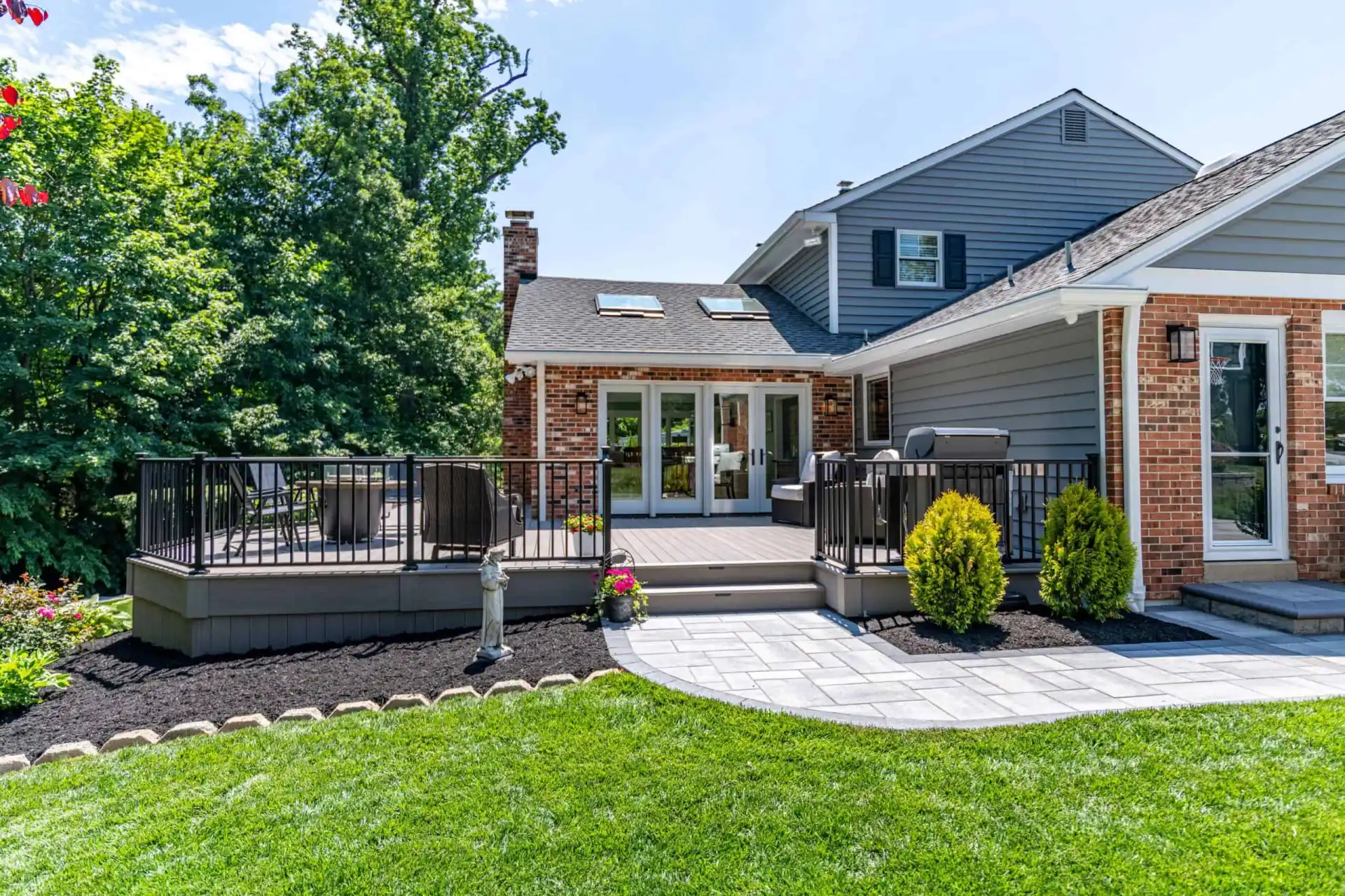 The backyard features a raised wooden deck with black railing, ideal for enjoying outdoor living in Delaware County, PA. Chairs and a table complete the setting. The house boasts brick and gray siding, while lush green grass and landscaped bushes enhance the space with trees in the background.