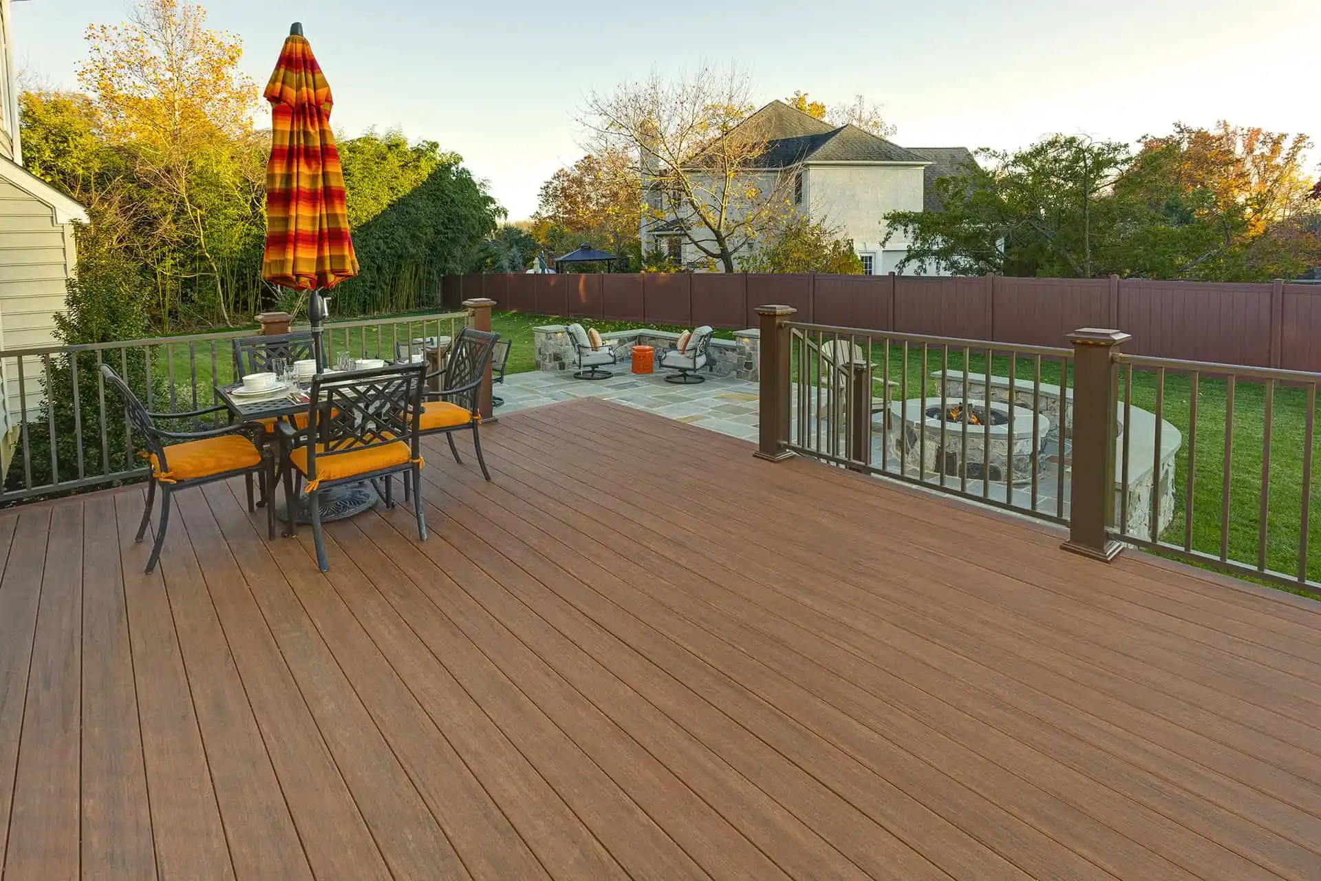 A spacious backyard deck with a table, chairs, and an umbrella, overlooking a fenced yard with a patio area, fire pit, and additional seating, surrounded by trees and a house in the background.