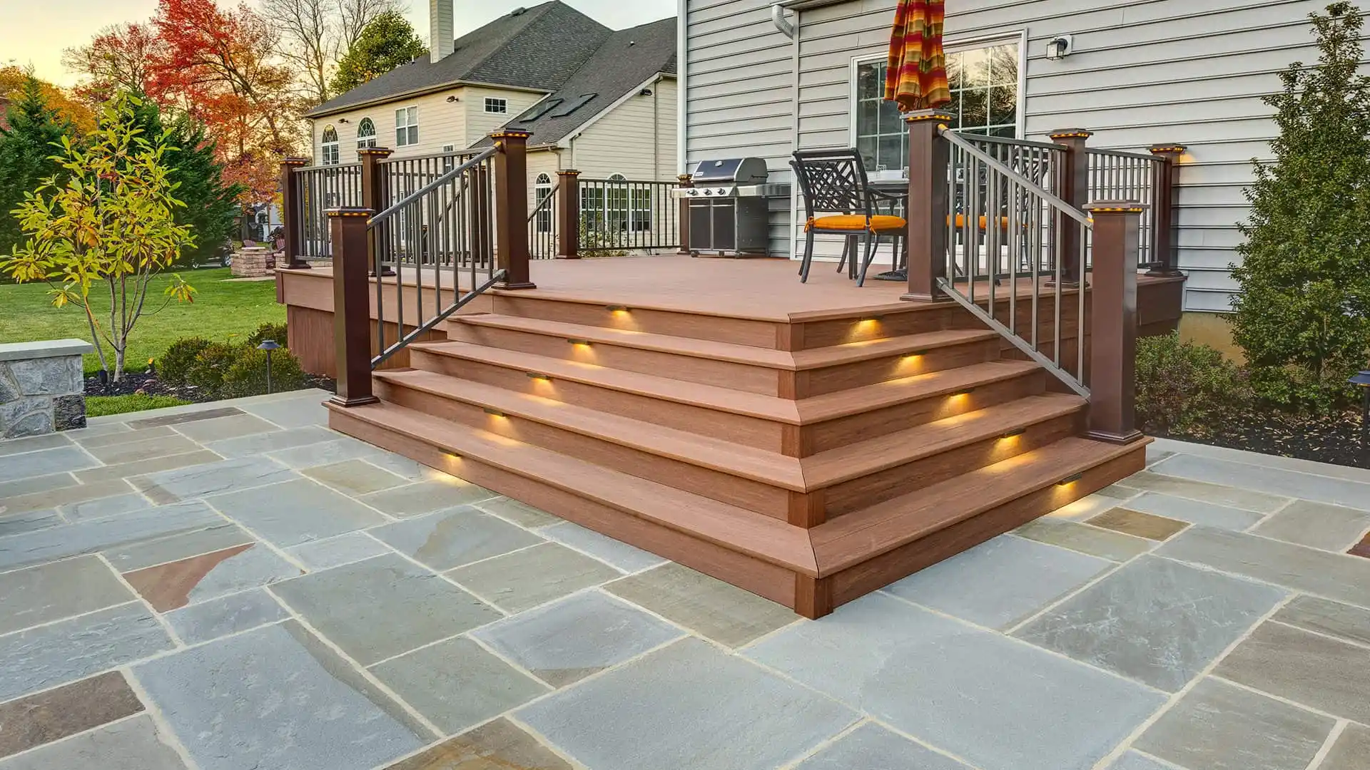 A wooden deck with illuminated steps leads to a house, featuring a grill and orange-cushioned patio chairs. Perfect for enjoying the outdoors in Delaware County, PA, it overlooks expert hardscaping and a grassy yard with trees and a glimpse of the neighboring house.