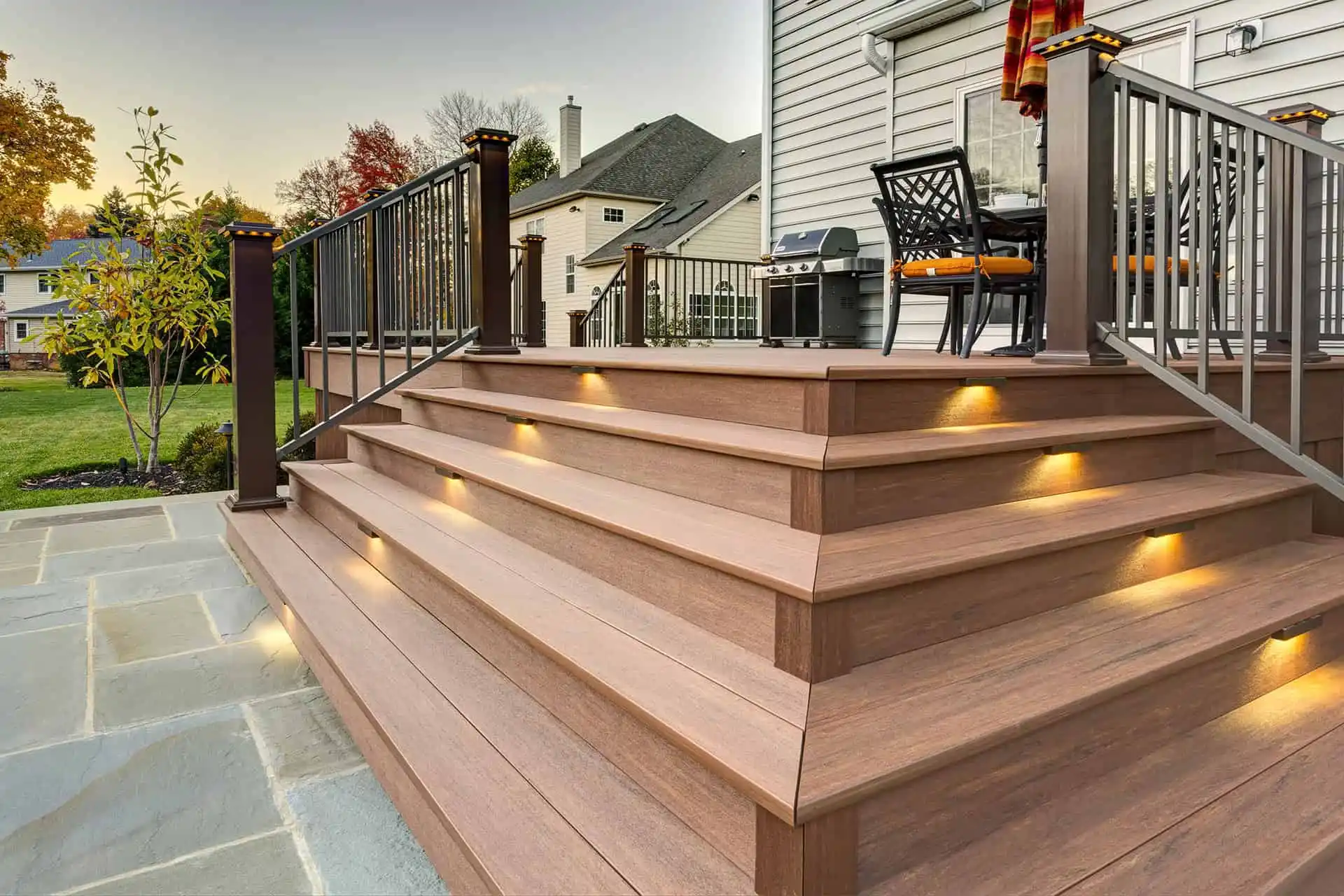 A modern wooden deck with wide steps, built-in step lighting, and metal railings, attached to a house. Outdoor furniture and a grill are visible on the deck, with a lawn and neighboring homes in the background.