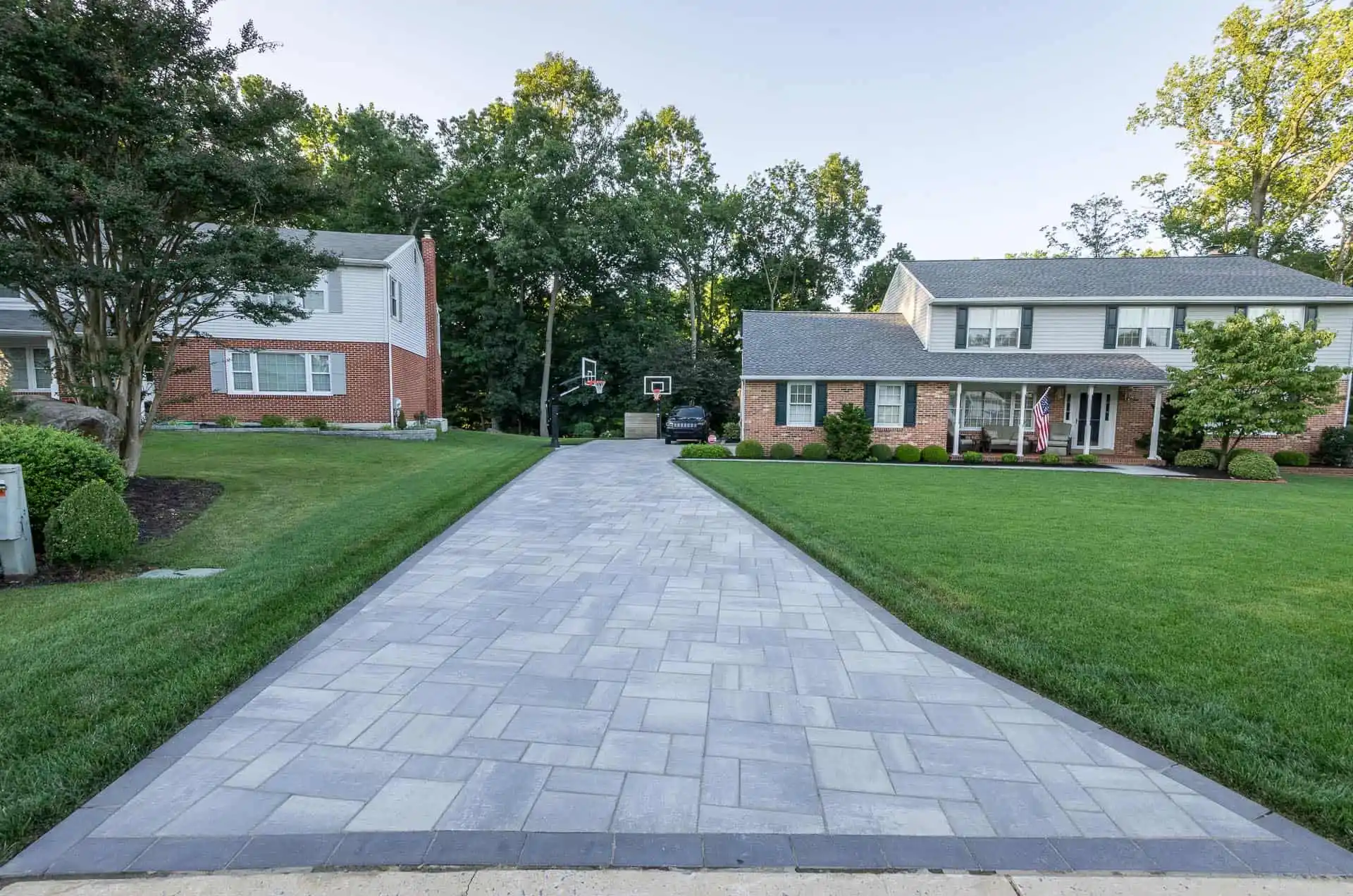 A newly paved stone driveway leads to a suburban two-story house with a front lawn, trees in the background, and a basketball hoop near the garage.