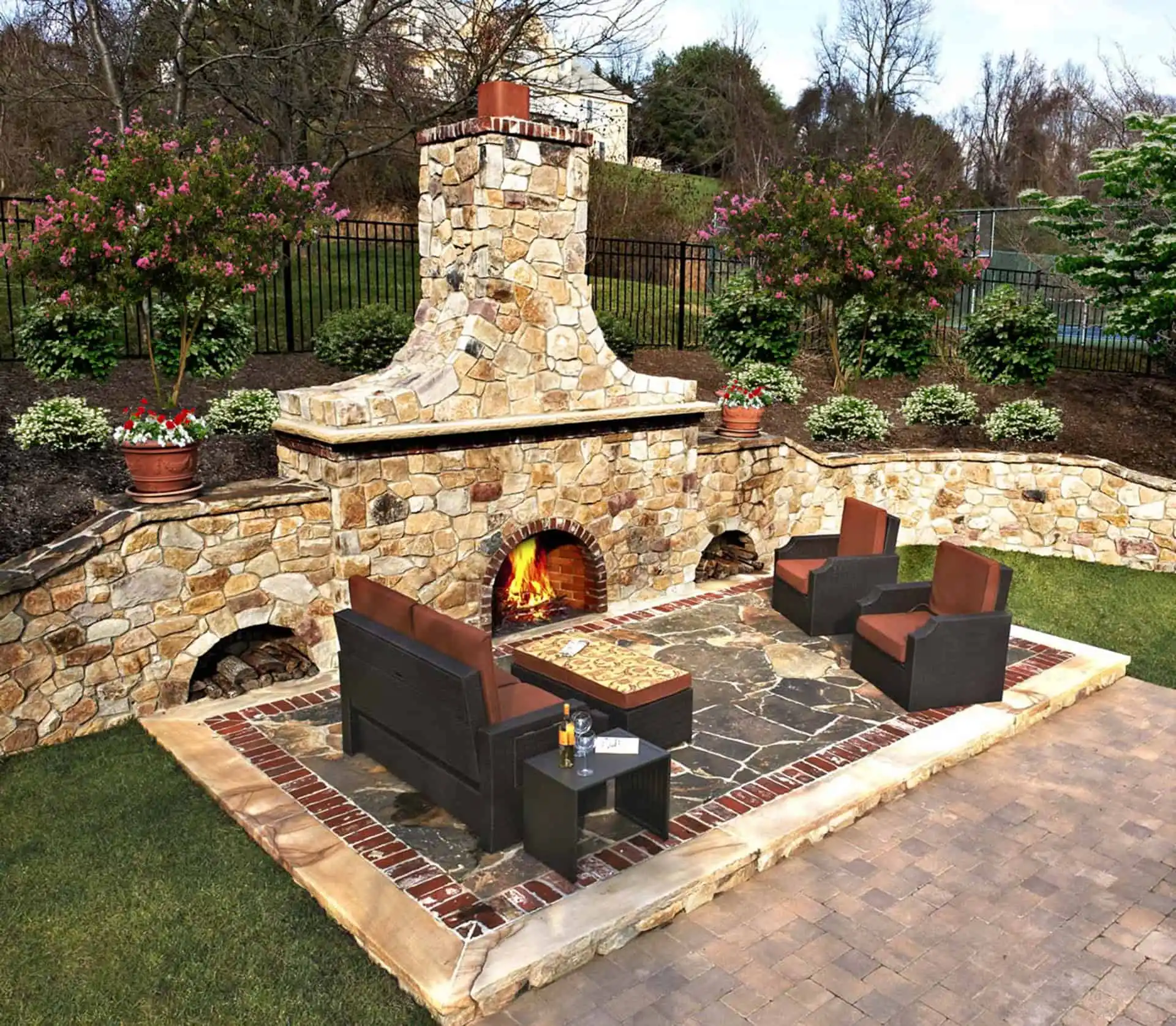 Outdoor patio with a stone fireplace, fire burning inside, surrounded by cushioned chairs and a bench, a table with drinks, and landscaped garden beds with blooming bushes and potted plants in the background.