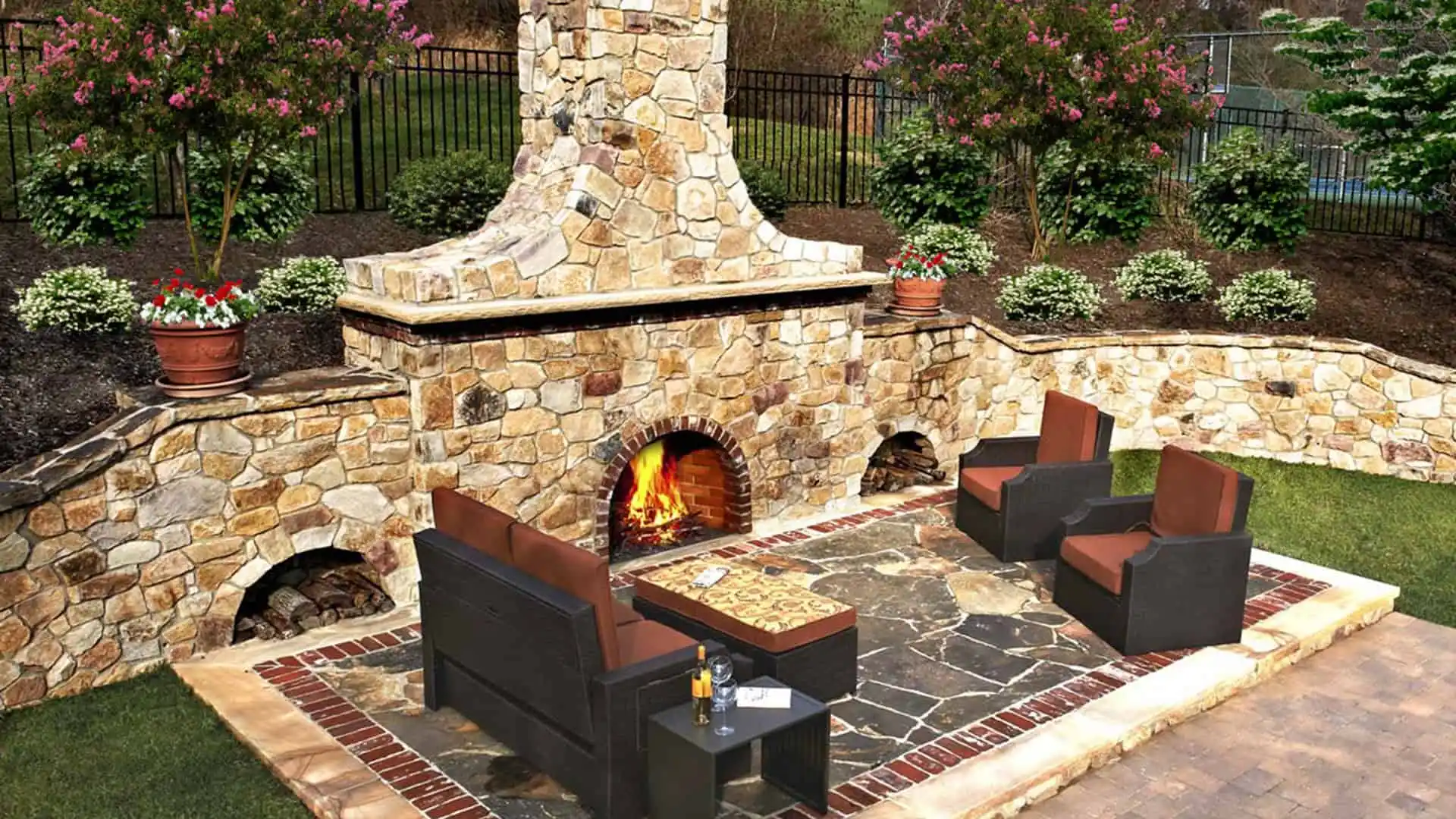 A cozy outdoor patio in Delaware County, PA, features a stone fireplace with four cushioned chairs around a table. The roaring fire adds warmth, while potted plants and shrubs enhance the charm. Ideal for enjoying outdoor living, thanks to expert hardscaping services in the area.