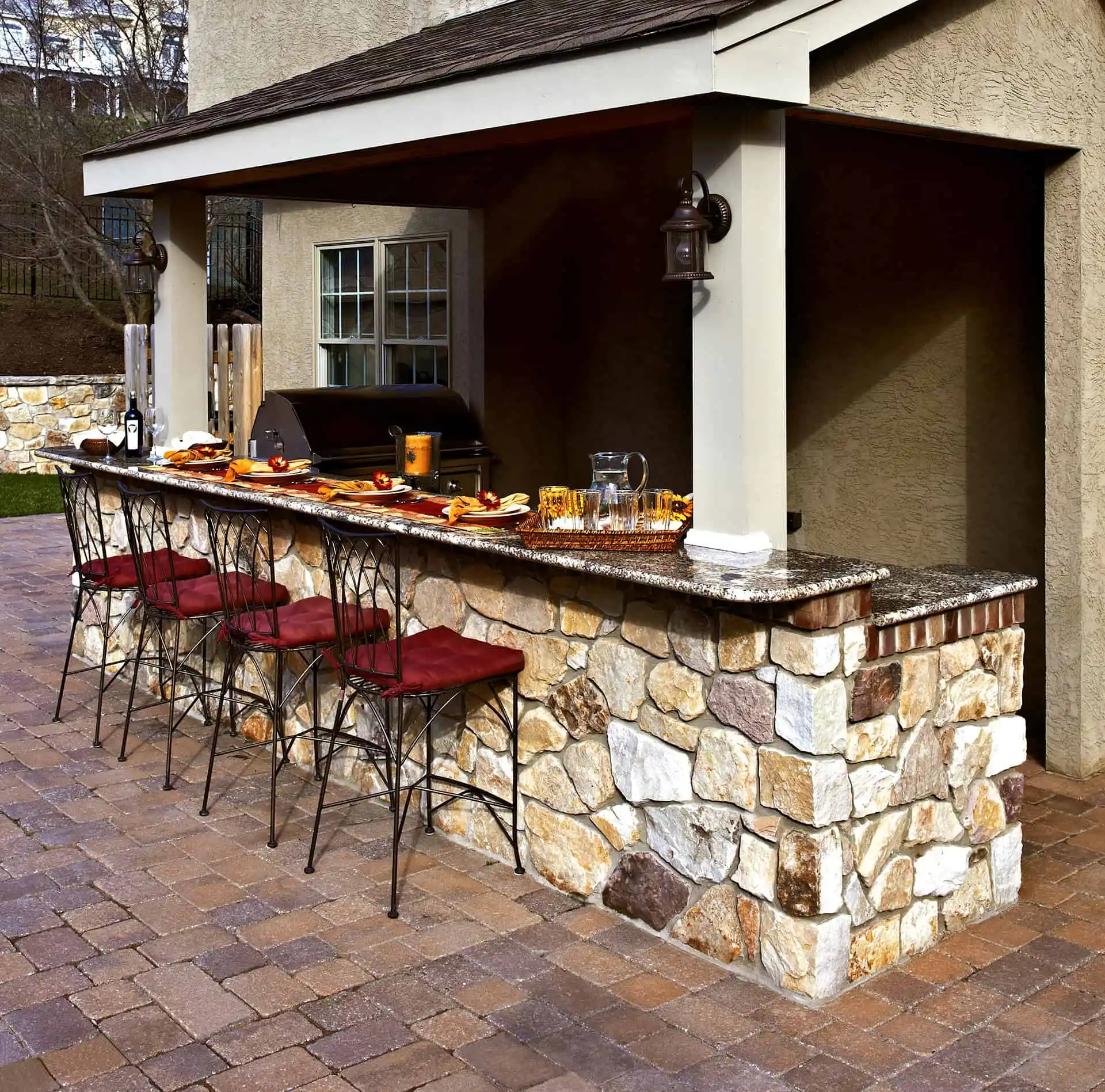 Outdoor kitchen with a stone bar, four metal barstools with red cushions, a countertop set with food and drinks, and a grill under a covered patio area. The floor is paved with bricks.