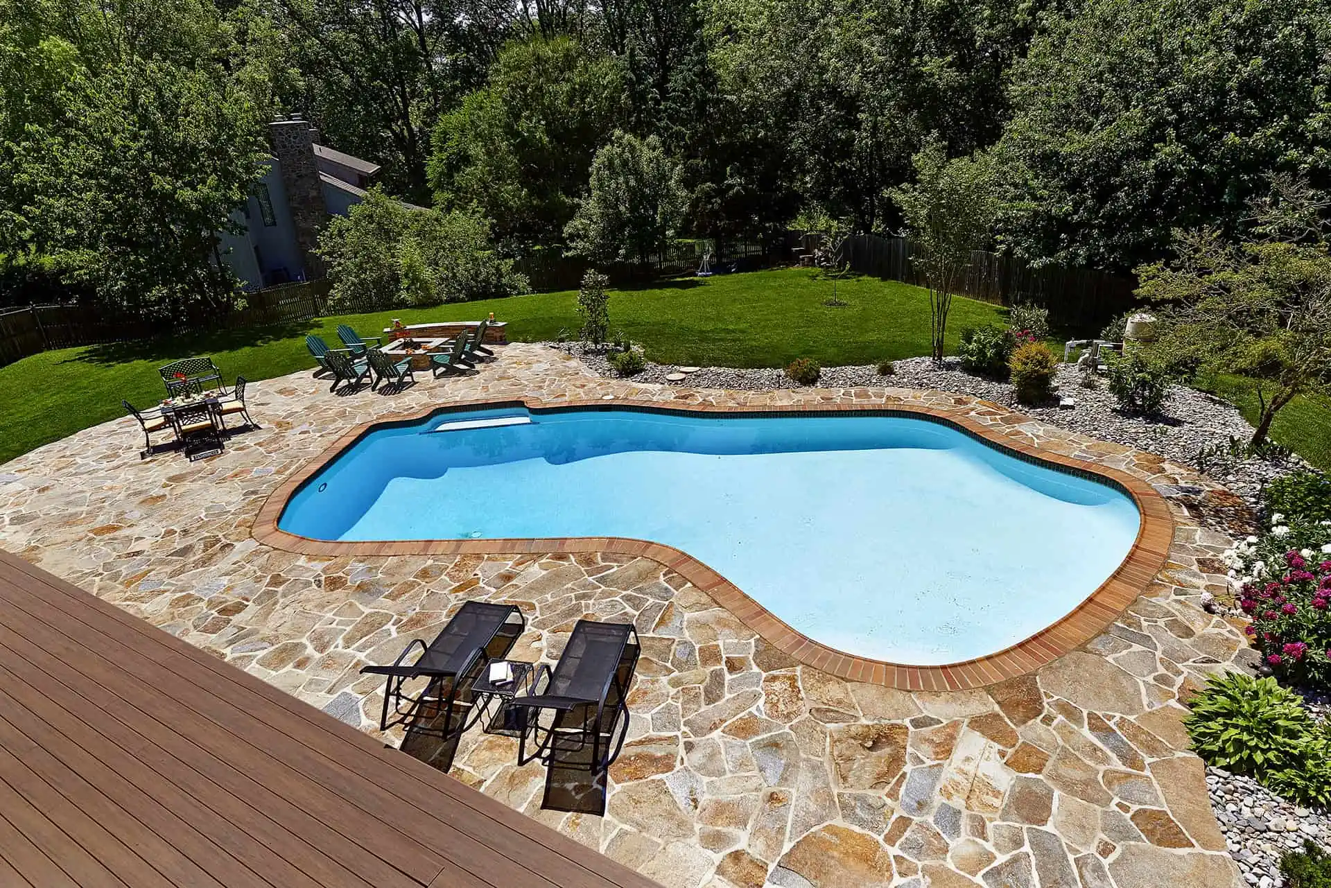 A backyard swimming pool with clear blue water, surrounded by a stone patio, lounge chairs, and green lawn with trees and shrubs in the background.