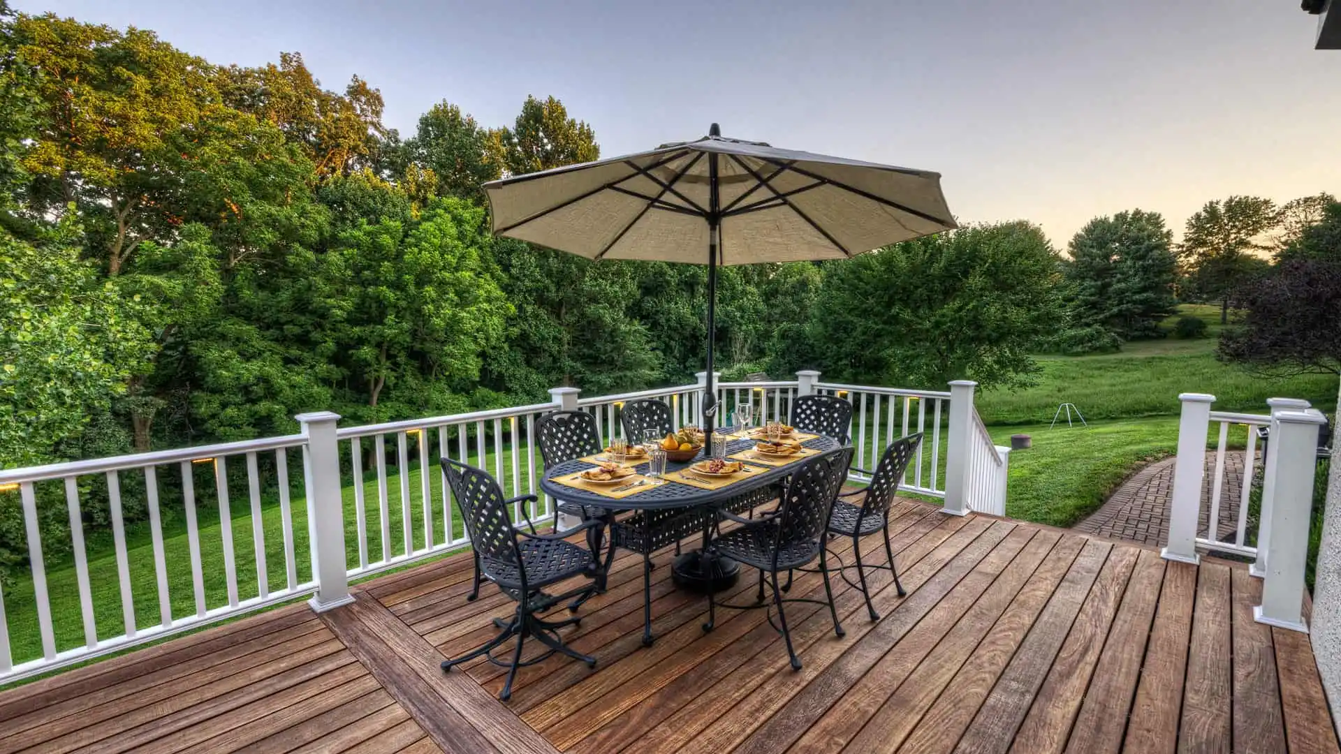 A wooden deck with a dining table set for six, featuring decorative plates and a large umbrella. Surrounded by lush green trees and grass under a clear sky, it’s an epitome of outdoor living in Delaware County, PA.