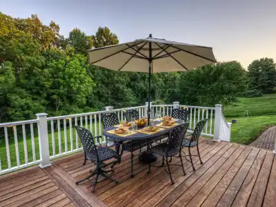 A wooden deck with a dining table set for six, featuring decorative plates and a large umbrella. Surrounded by lush green trees and grass under a clear sky, it’s an epitome of outdoor living in Delaware County, PA.
