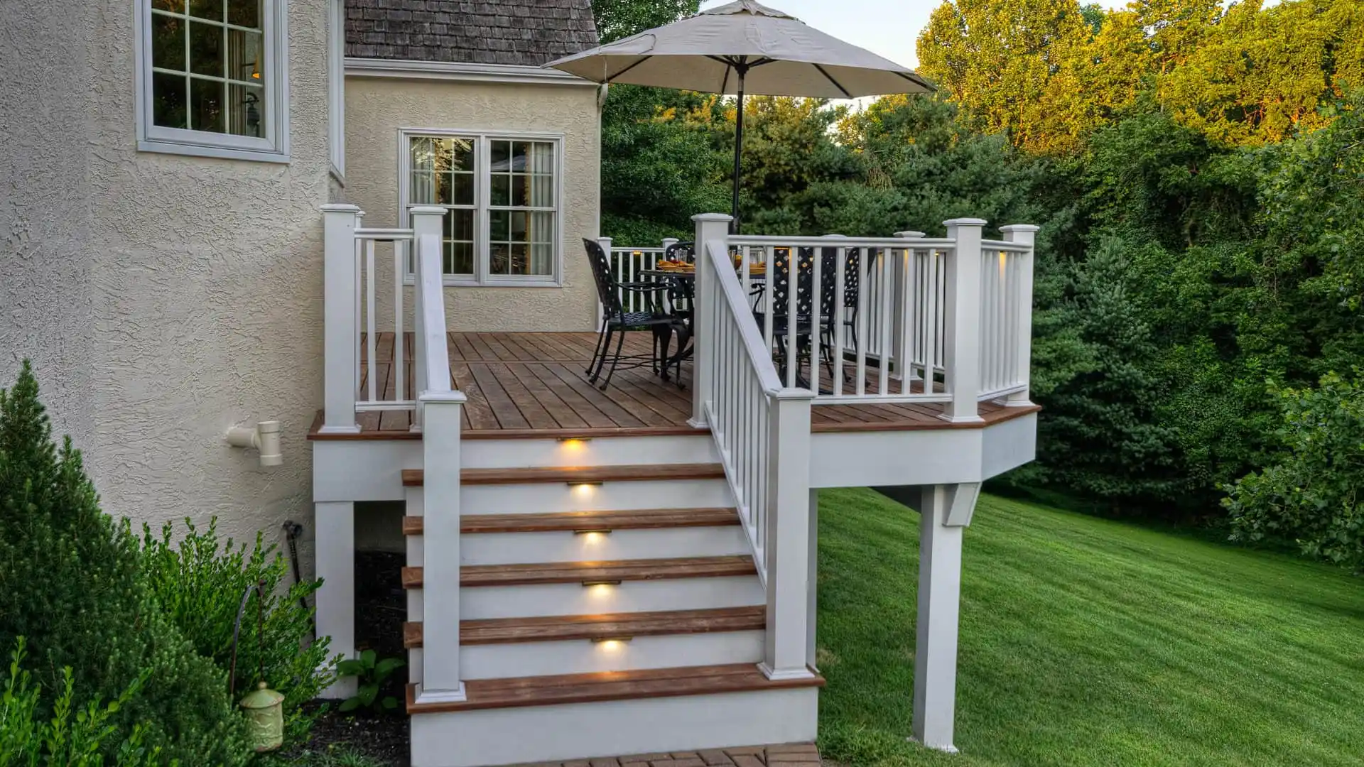Elevated wooden deck with stairs and built-in step lights, perfect for outdoor living in Delaware County, PA. It features a table, chairs, and large umbrella. The light-colored house's deck overlooks a lush green yard surrounded by trees.