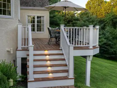 Elevated wooden deck with stairs and built-in step lights, perfect for outdoor living in Delaware County, PA. It features a table, chairs, and large umbrella. The light-colored house's deck overlooks a lush green yard surrounded by trees.