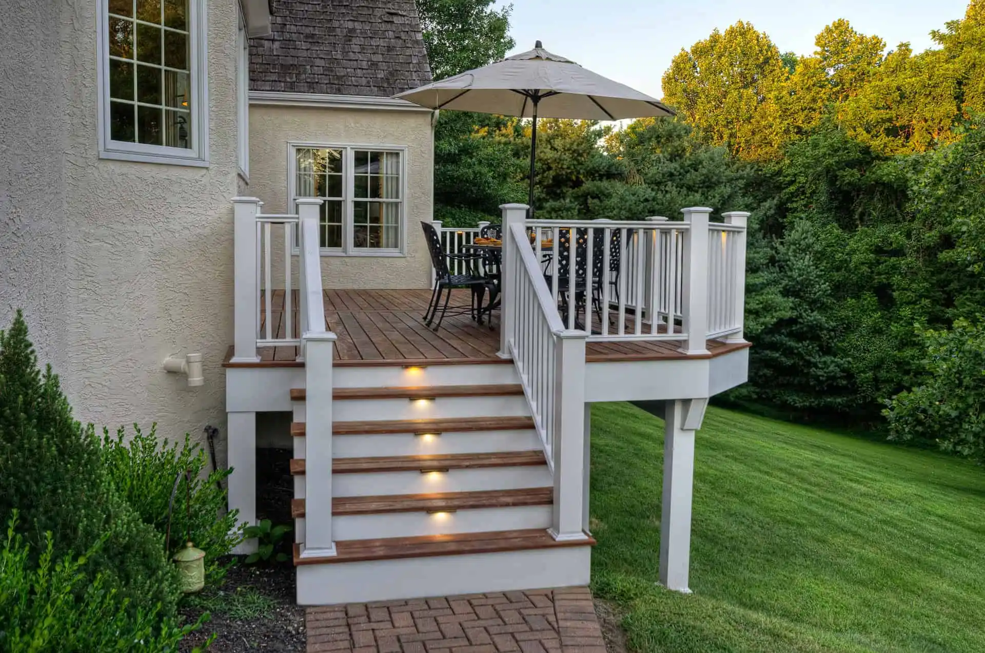 A wooden deck with white railings and brown trim features stairs with built-in lights, outdoor dining furniture, and a large umbrella, overlooking a green lawn and trees in the background.