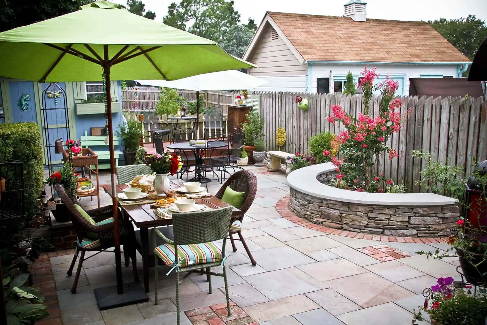 A cozy backyard patio with stone paving, a dining table set for a meal under a green umbrella, colorful chairs, flowering plants, and a curved stone bench beside a wooden fence. A house and shed are in the background.