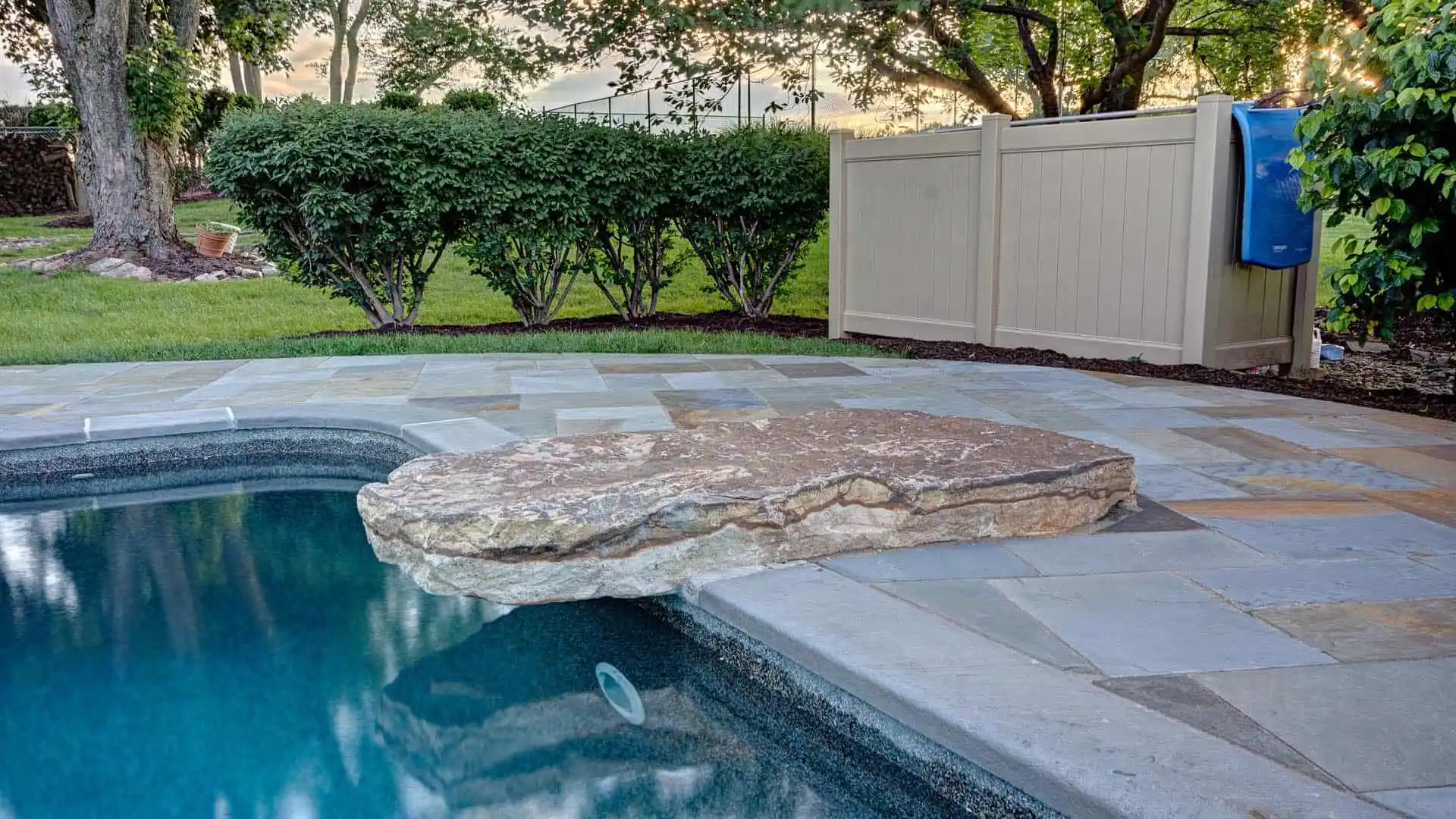 A backyard pool features a large, flat stone extending over its edge, surrounded by a patio of grand stone tiles—a perfect example of outdoor living in Delaware County, PA. Bushes and trees frame the scene, with a beige privacy screen providing seclusion on the right.