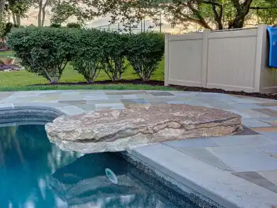 A backyard pool features a large, flat stone extending over its edge, surrounded by a patio of grand stone tiles—a perfect example of outdoor living in Delaware County, PA. Bushes and trees frame the scene, with a beige privacy screen providing seclusion on the right.