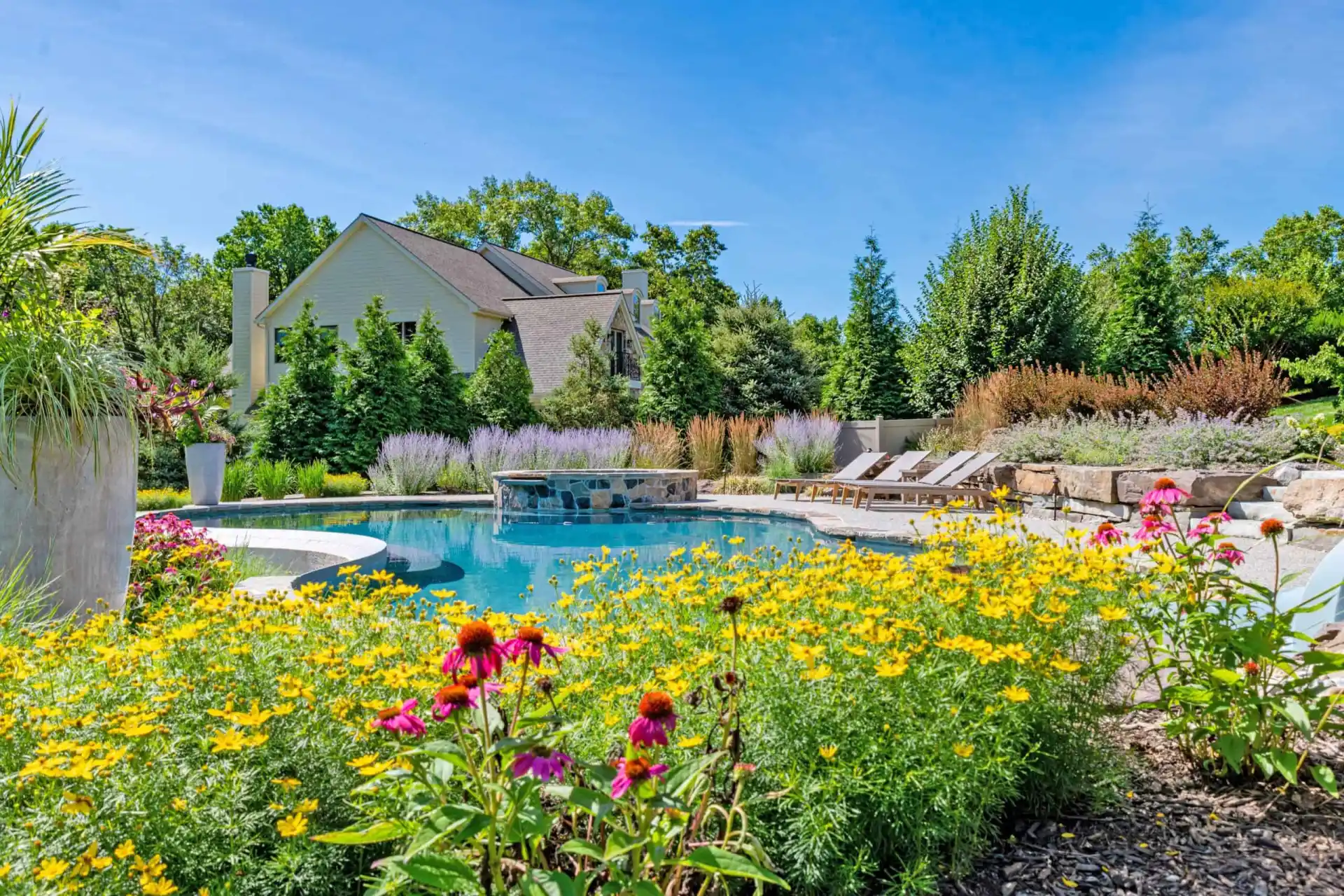 A backyard pool surrounded by vibrant flowers and greenery under a clear blue sky showcases the artistry of outdoor living Delaware County PA. Lounge chairs are positioned on one side, while a house with a beige exterior stands framed by tall trees in the background.